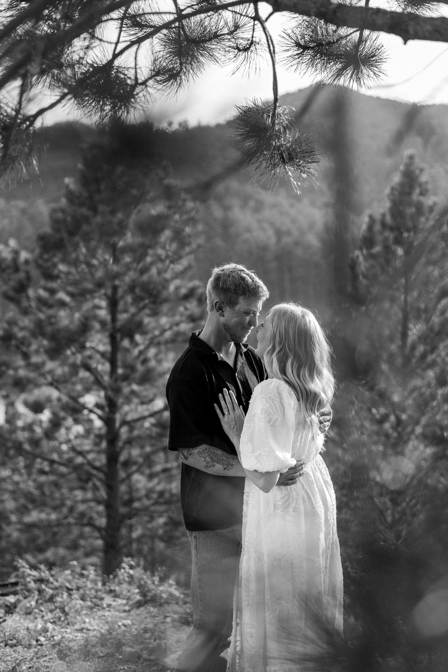 Romantic black and white portrait of a couple embracing under pine trees in a mountain setting.