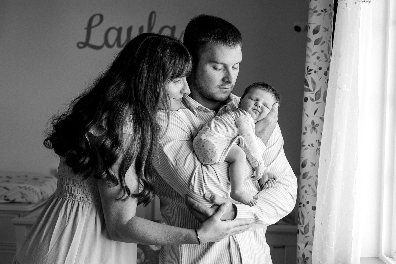 A tender black and white portrait of parents comforting their crying newborn baby by a window.