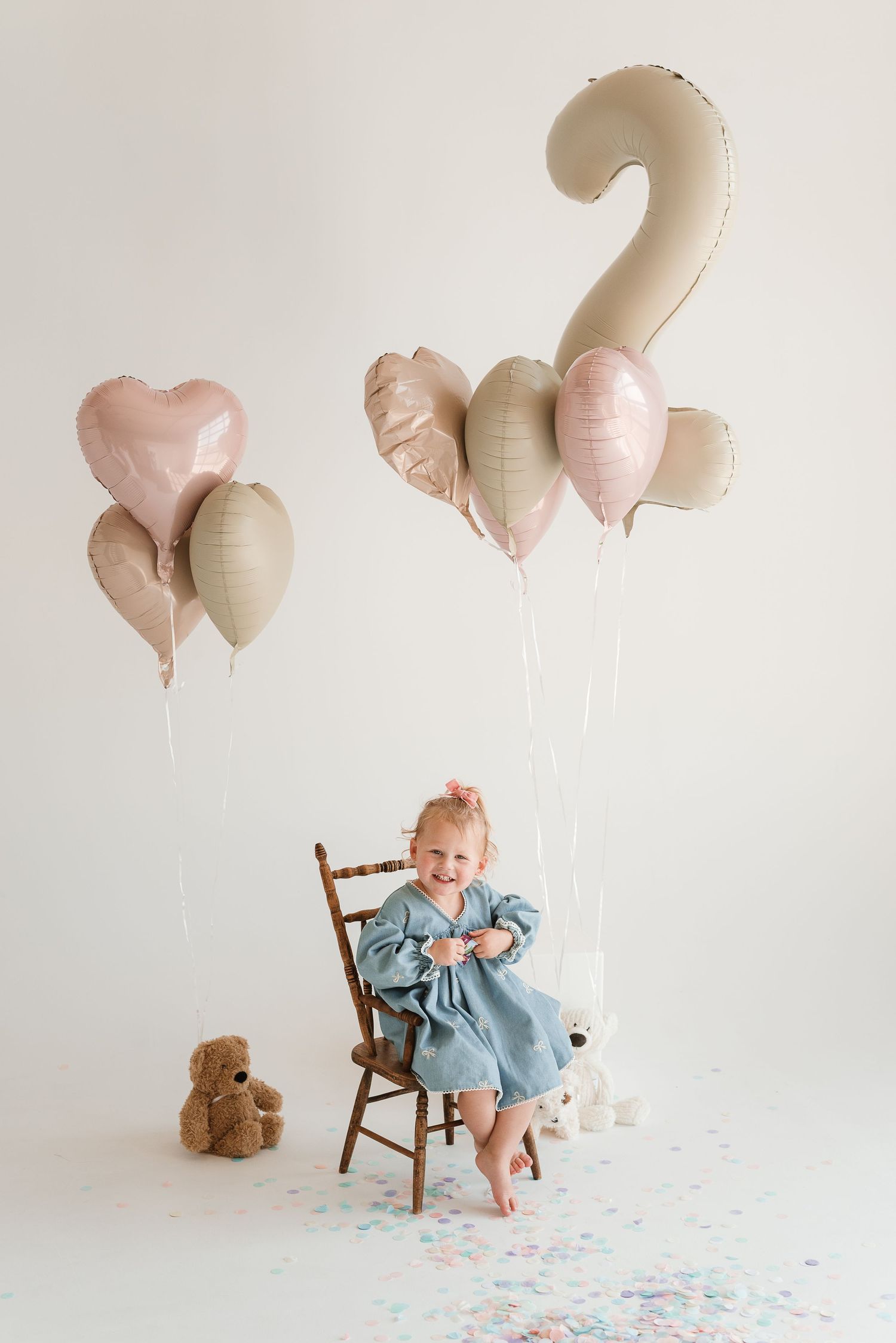 Birthday celebration scene with child in blue dress seated on wooden chair with pink and beige balloons.