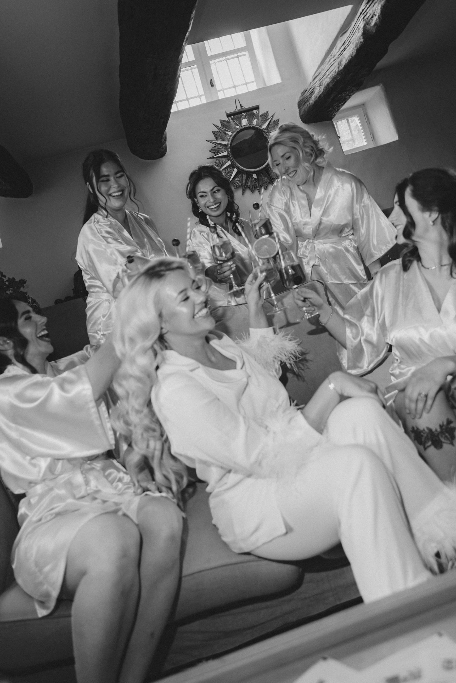 Black and white photo showing bridesmaids in white robes laughing and celebrating together on a couch.