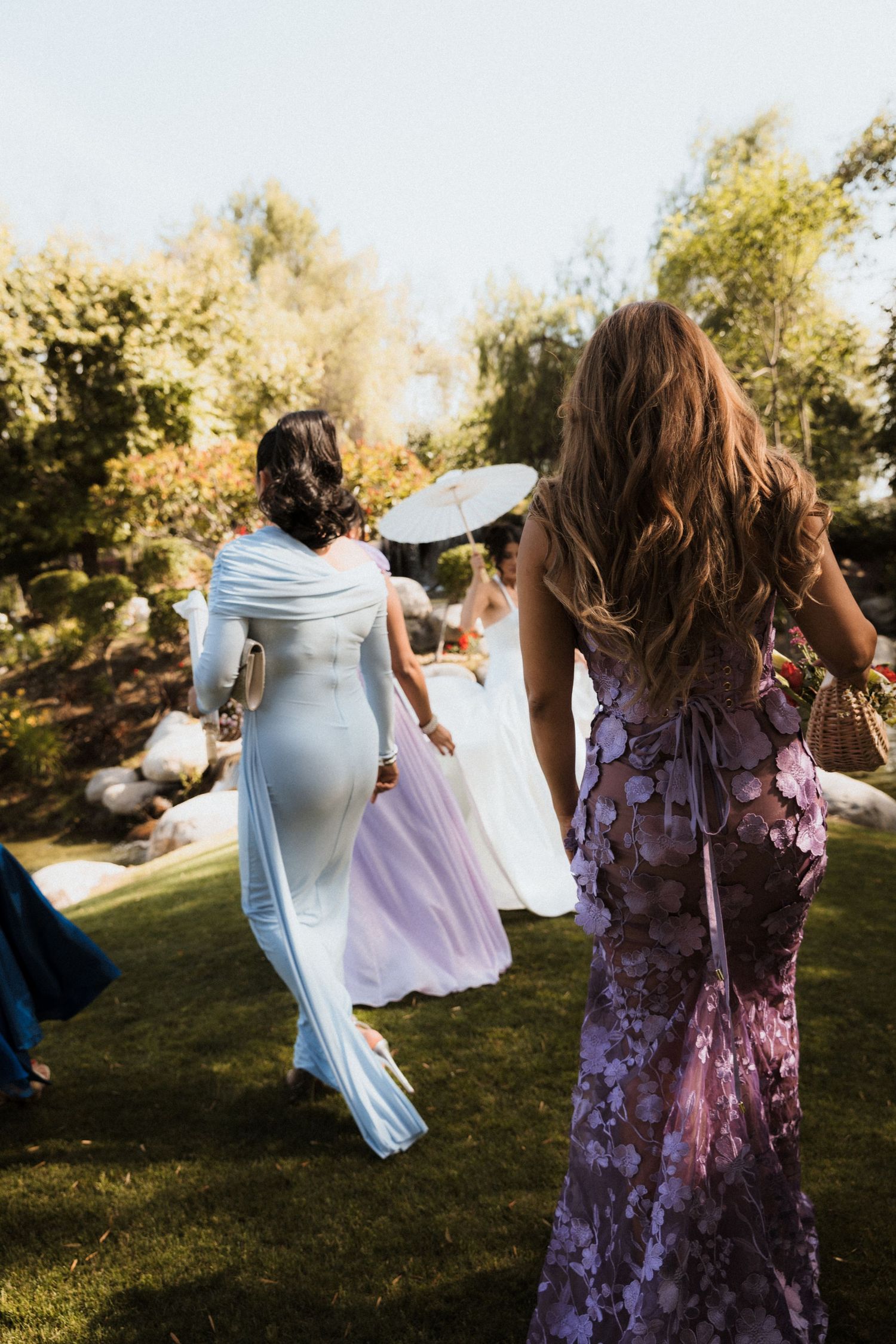 People in purple and light colored dresses dance and twirl together in a sunlit outdoor garden setting.