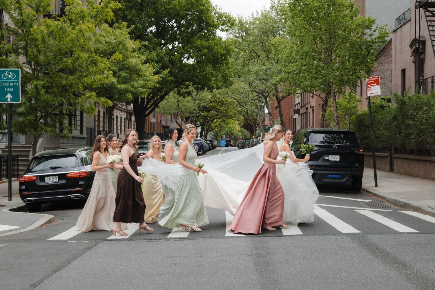 Bridal party in colorful gowns crossing a tree-lined street, bride's white dress flowing, urban wedding photo.