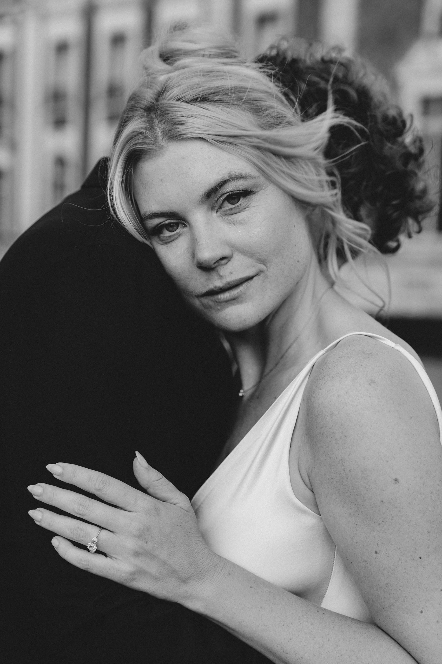 Black and white portrait of a blonde bride in white dress hugging groom, showcasing engagement ring.