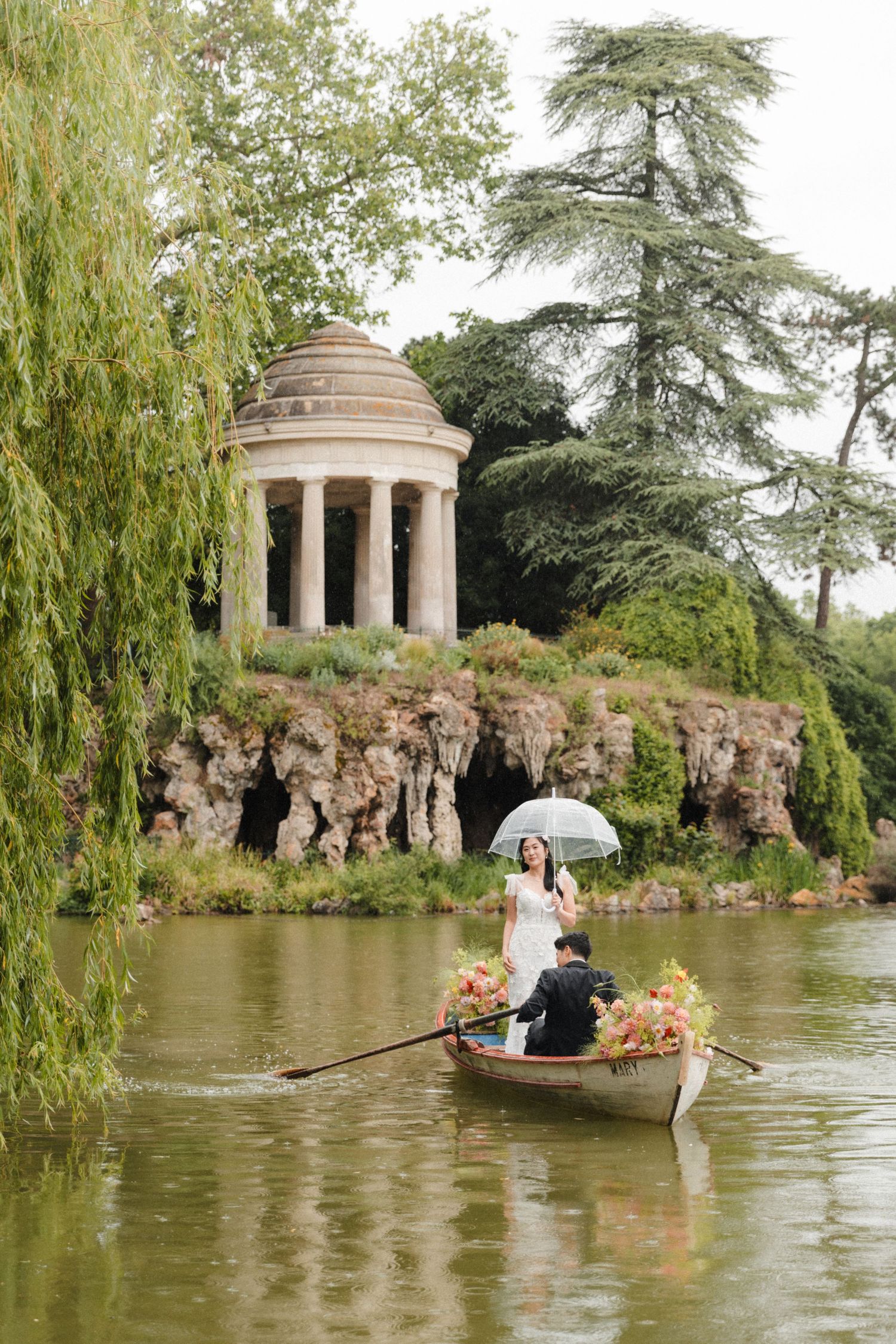Couple in a flower-filled rowboat on a lake with a classical temple and rocky grotto backdrop.