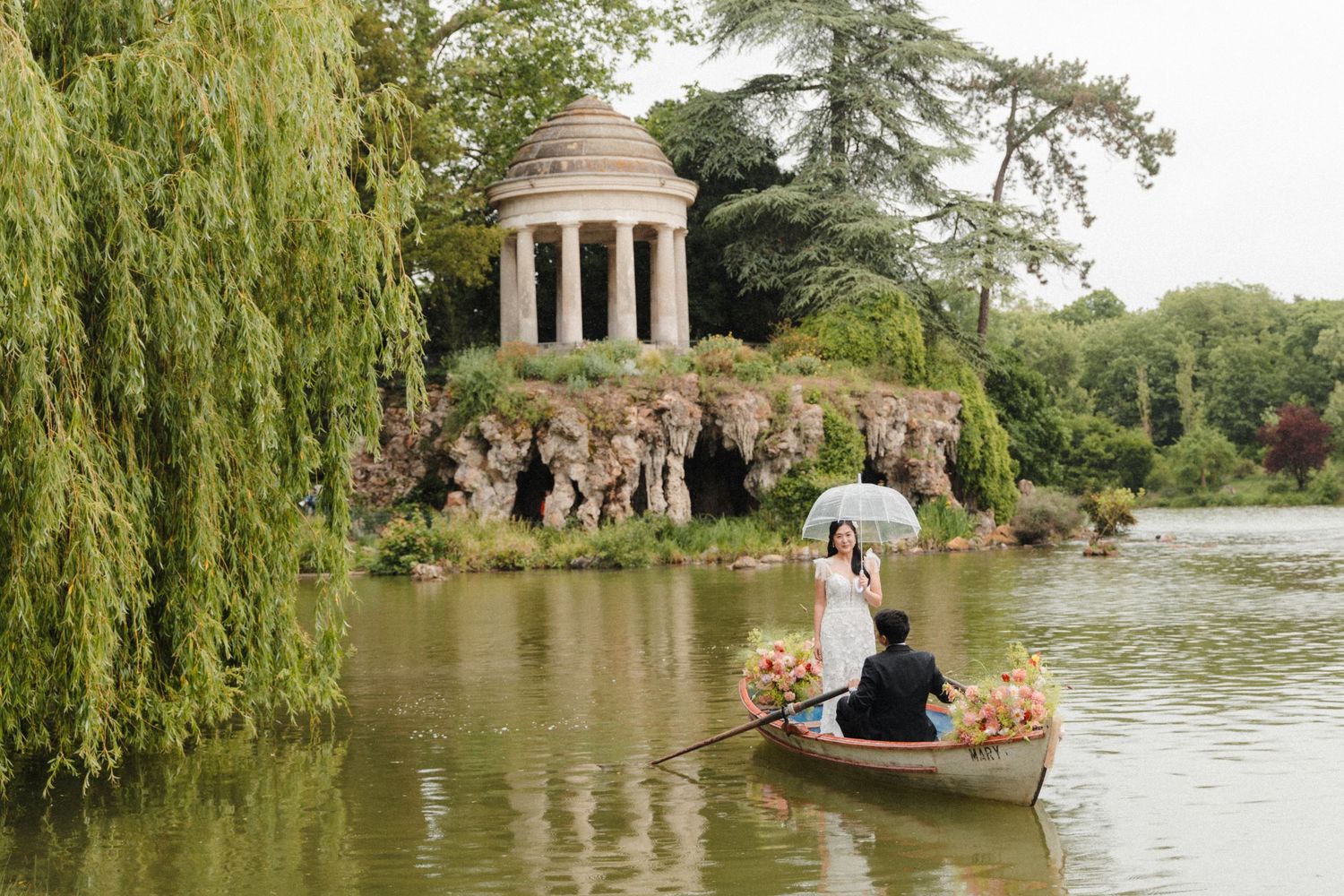 Couple in a rowboat on a serene lake with a classical temple and weeping willows at Bois de Vincennes, Paris.