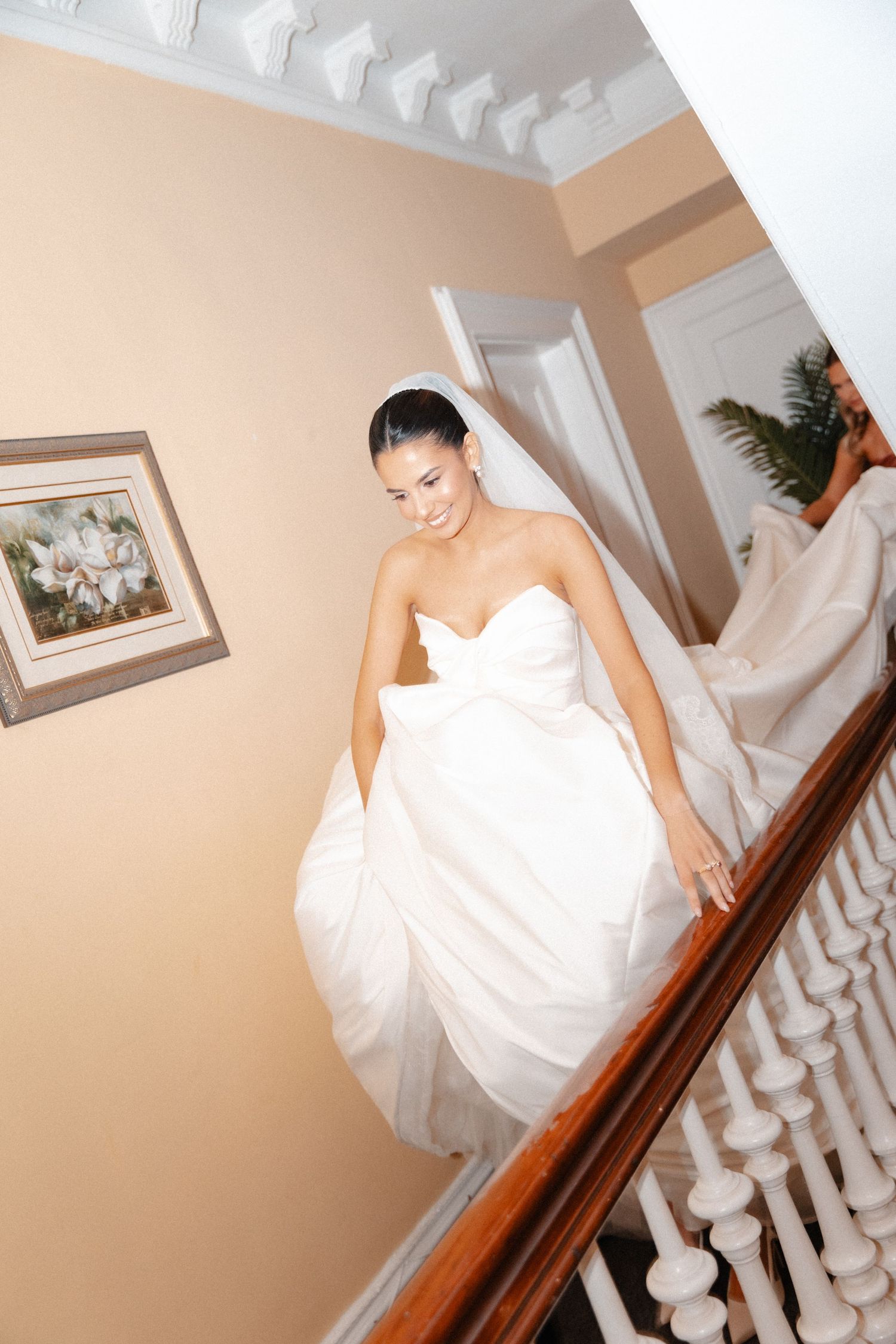 A smiling bride in a strapless white wedding gown walks down a staircase with a wooden banister.
