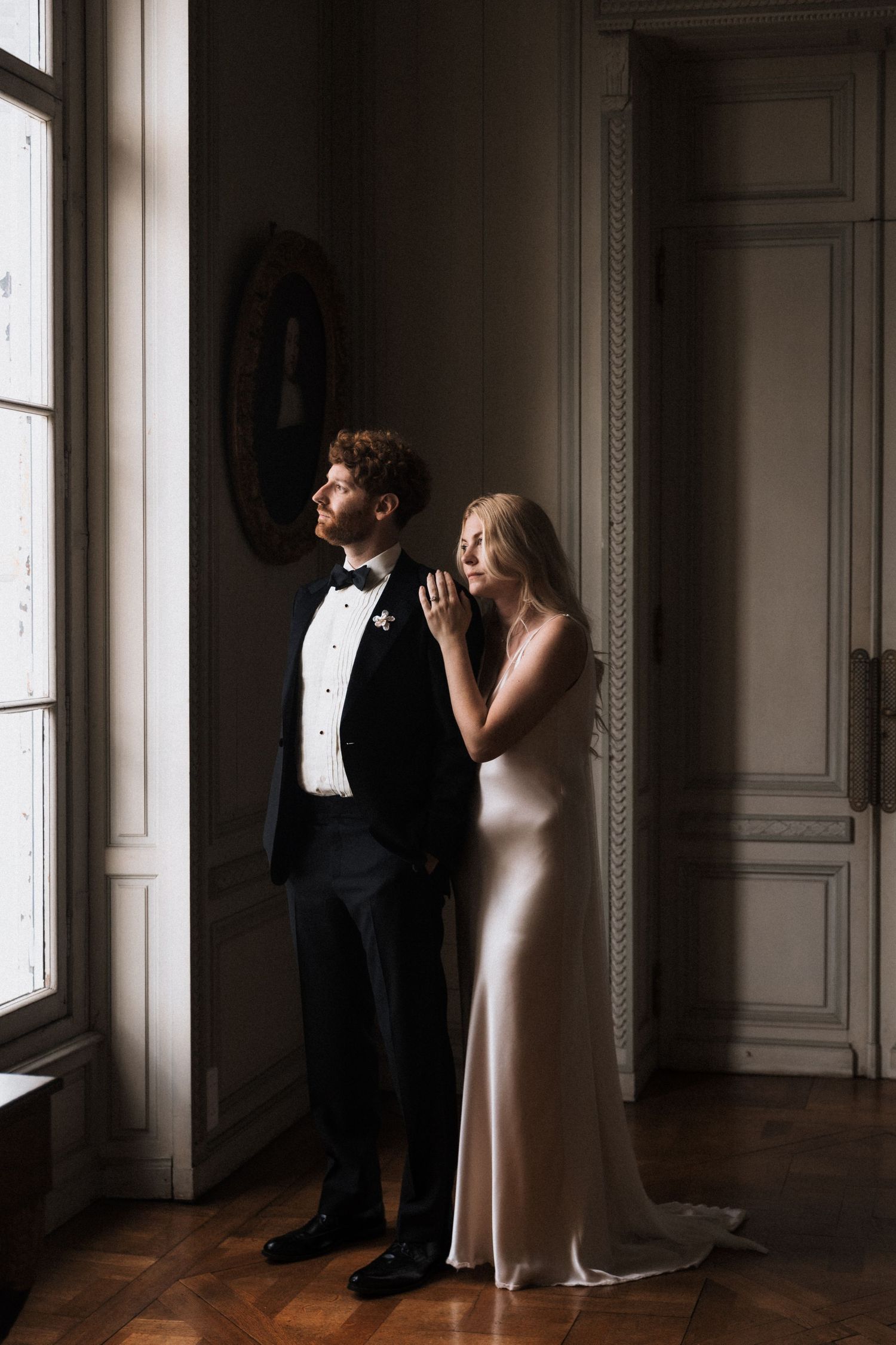 Elegant bride in silk gown and groom in tuxedo standing by window of french caslte