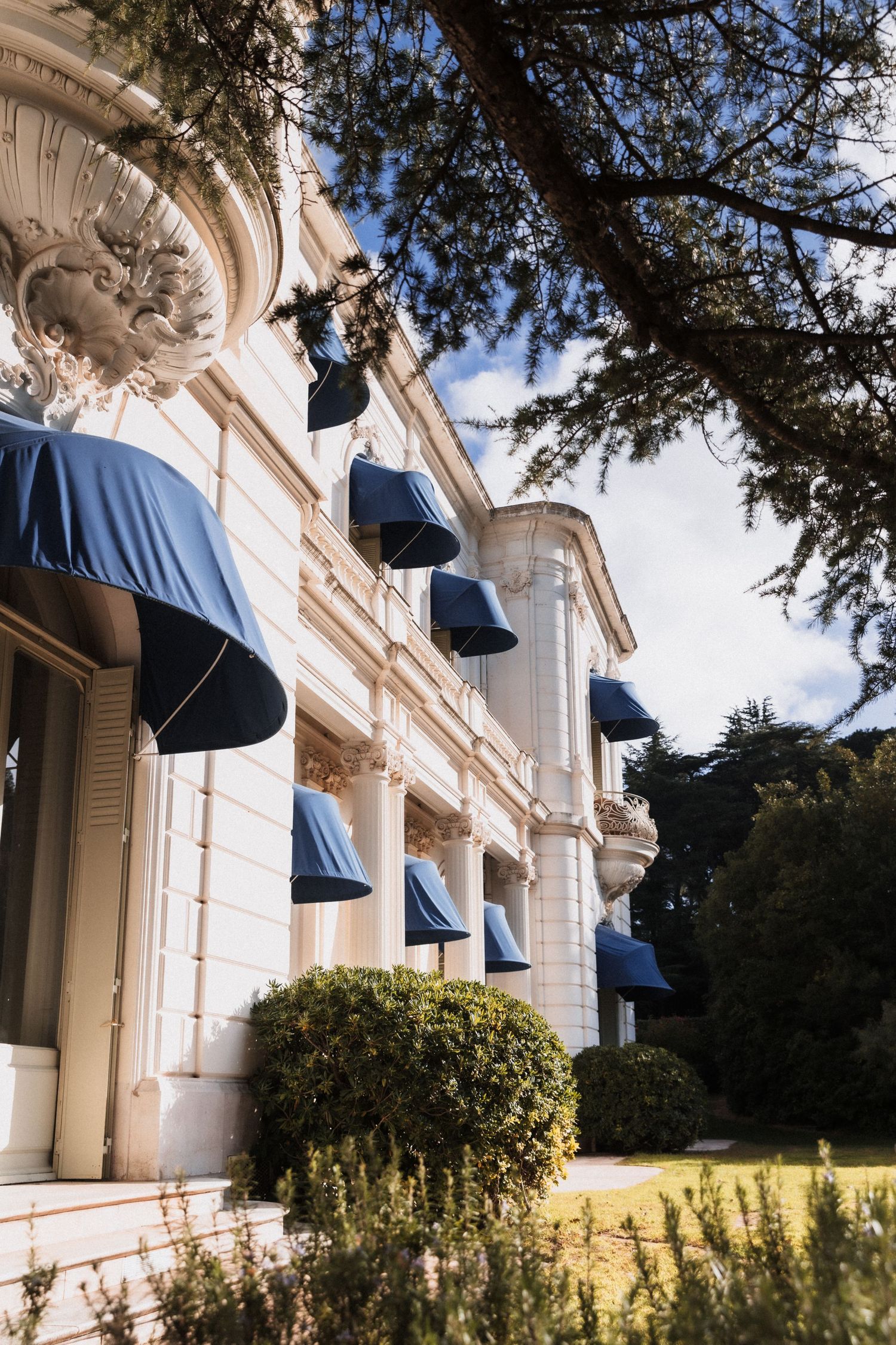 Villa Rocabella - Elegant white neoclassical building facade with blue awnings, ornate columns, and lush green shrubs