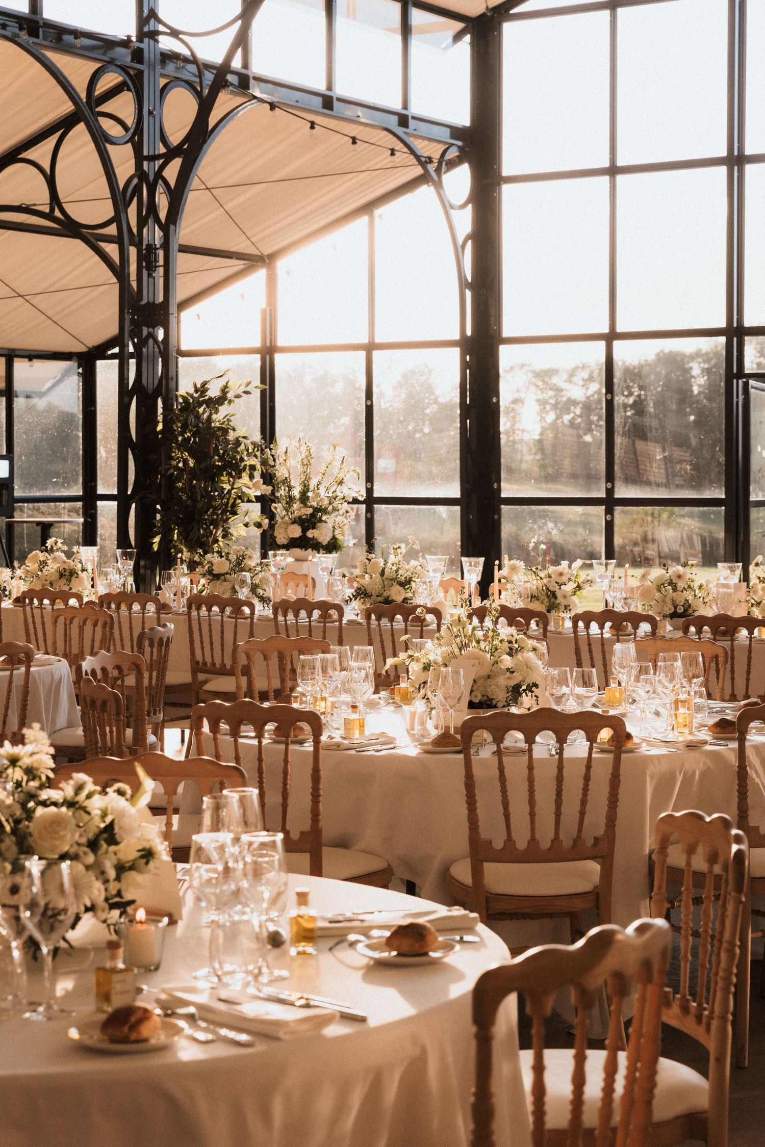 Elegant wedding reception venue with round tables set with white linens and floral centerpieces in a glass-walled space.
