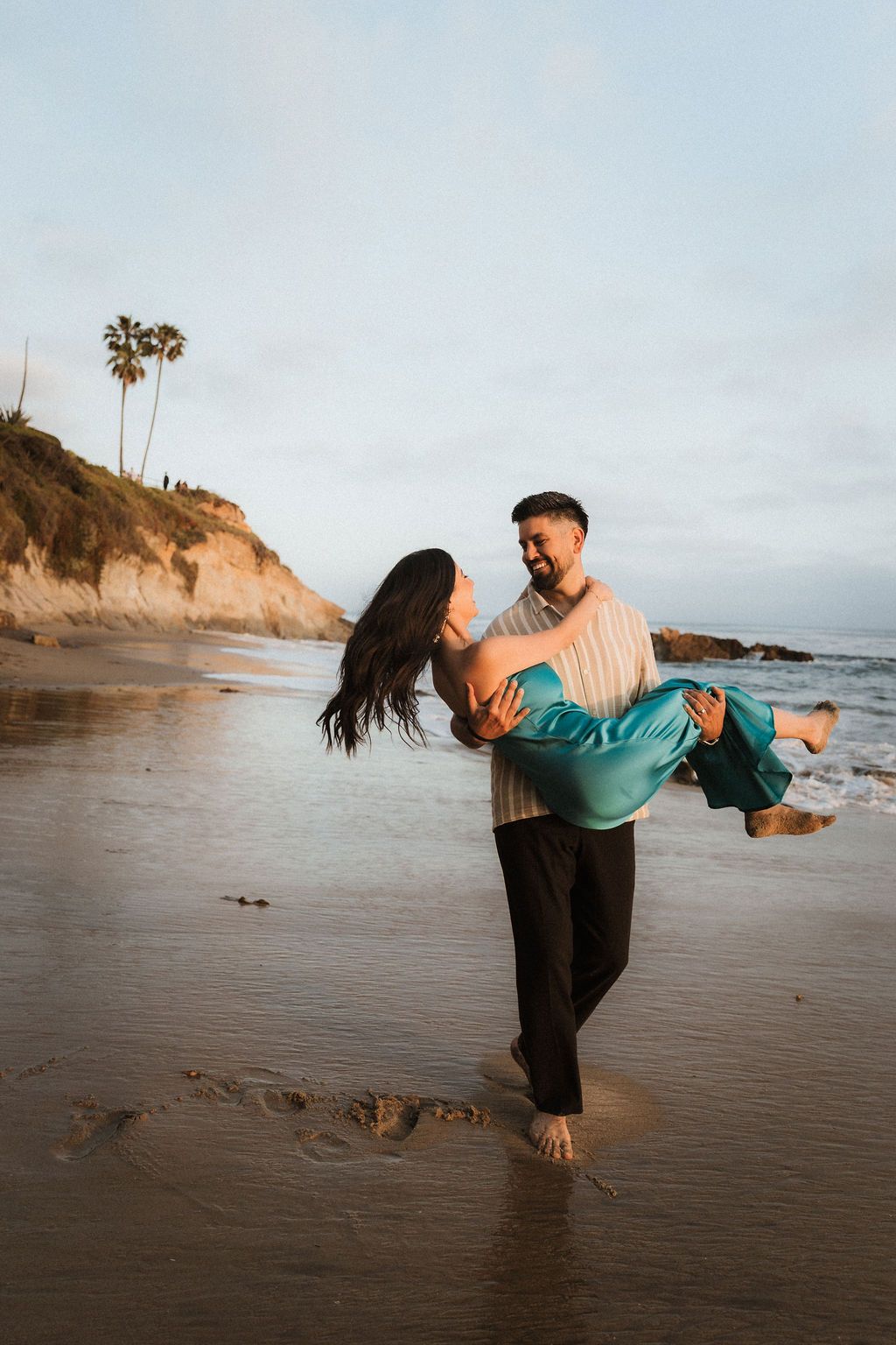 Editorial Laguna Beach engagement: Couple embracing at sunset with palm trees and rugged cliffs. Cinematic destination session