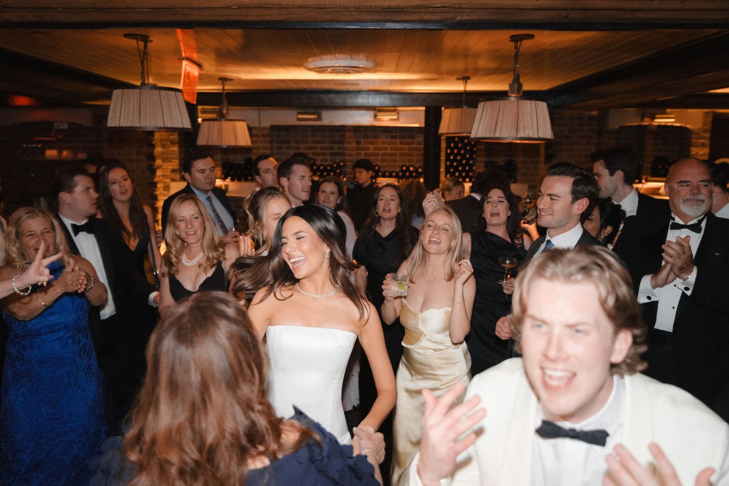 Bride in white strapless gown laughs with guests on wedding reception dance floor in elegant venue.