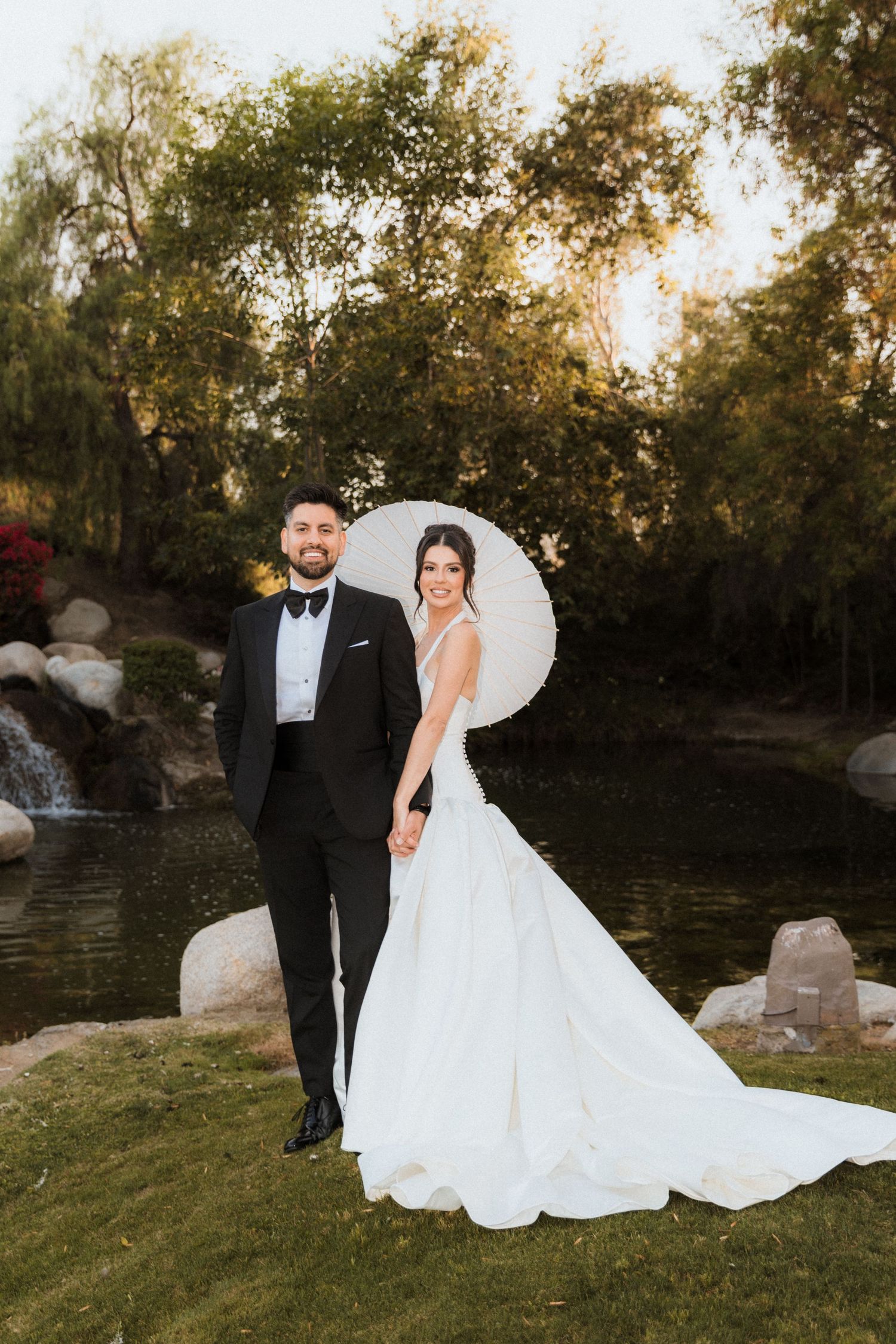 Bride and groom pose by a serene pond in a lush garden, she holds a white parasol wearing a flowing gown.
