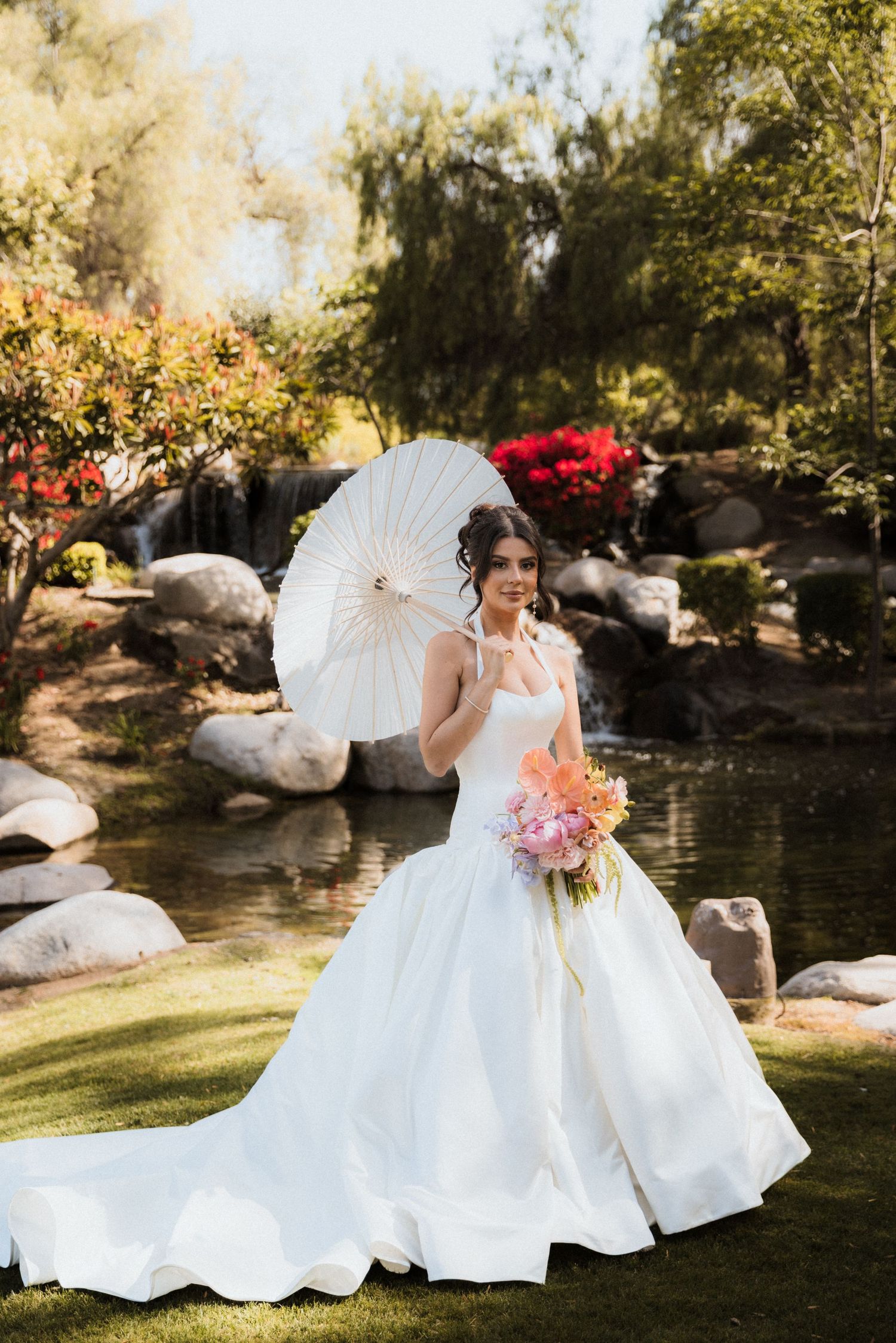 Bride in white ball gown holding parasol and floral bouquet by garden waterfall.