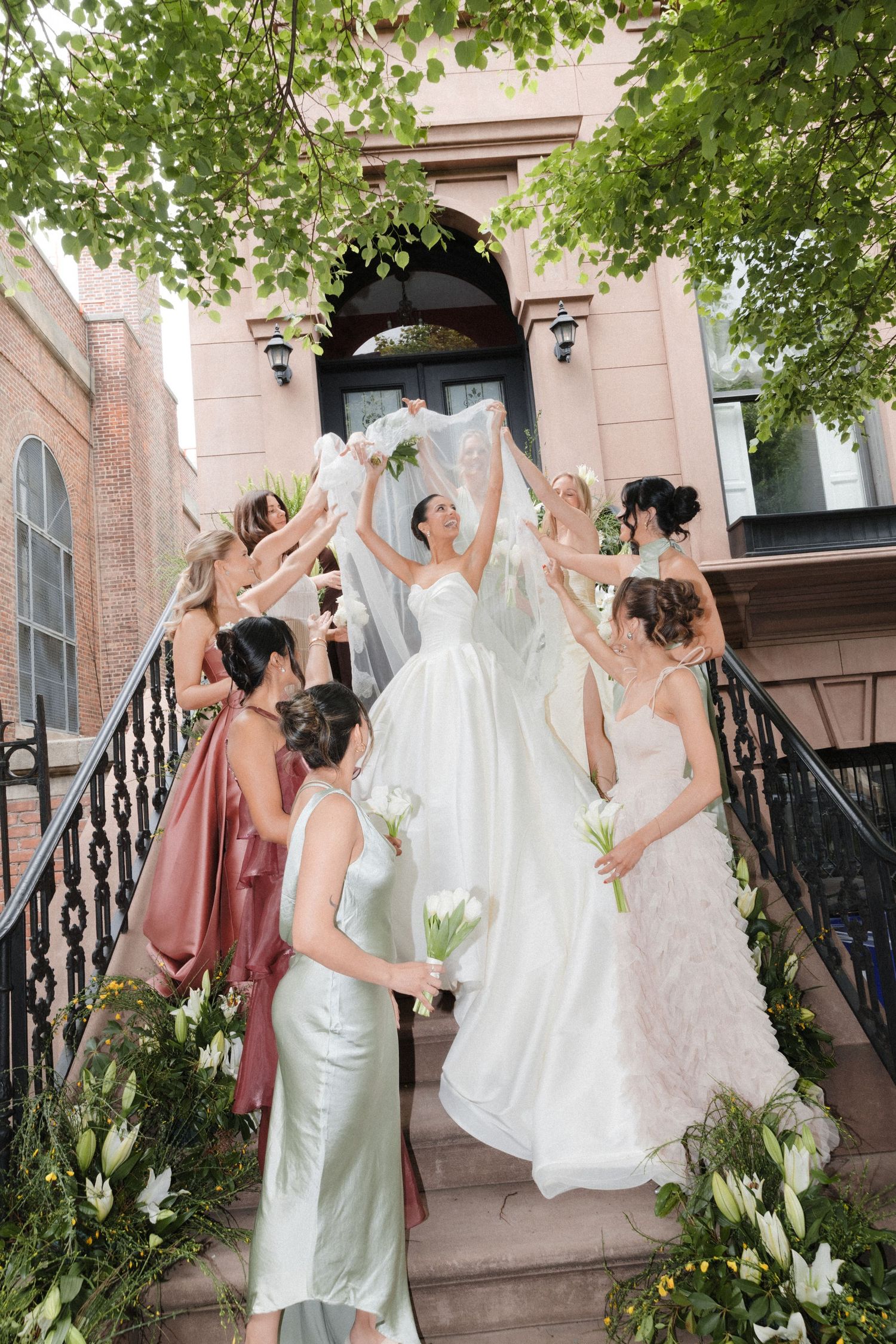 Bride in white gown celebrates with bridesmaids in colorful dresses on decorated brownstone steps.