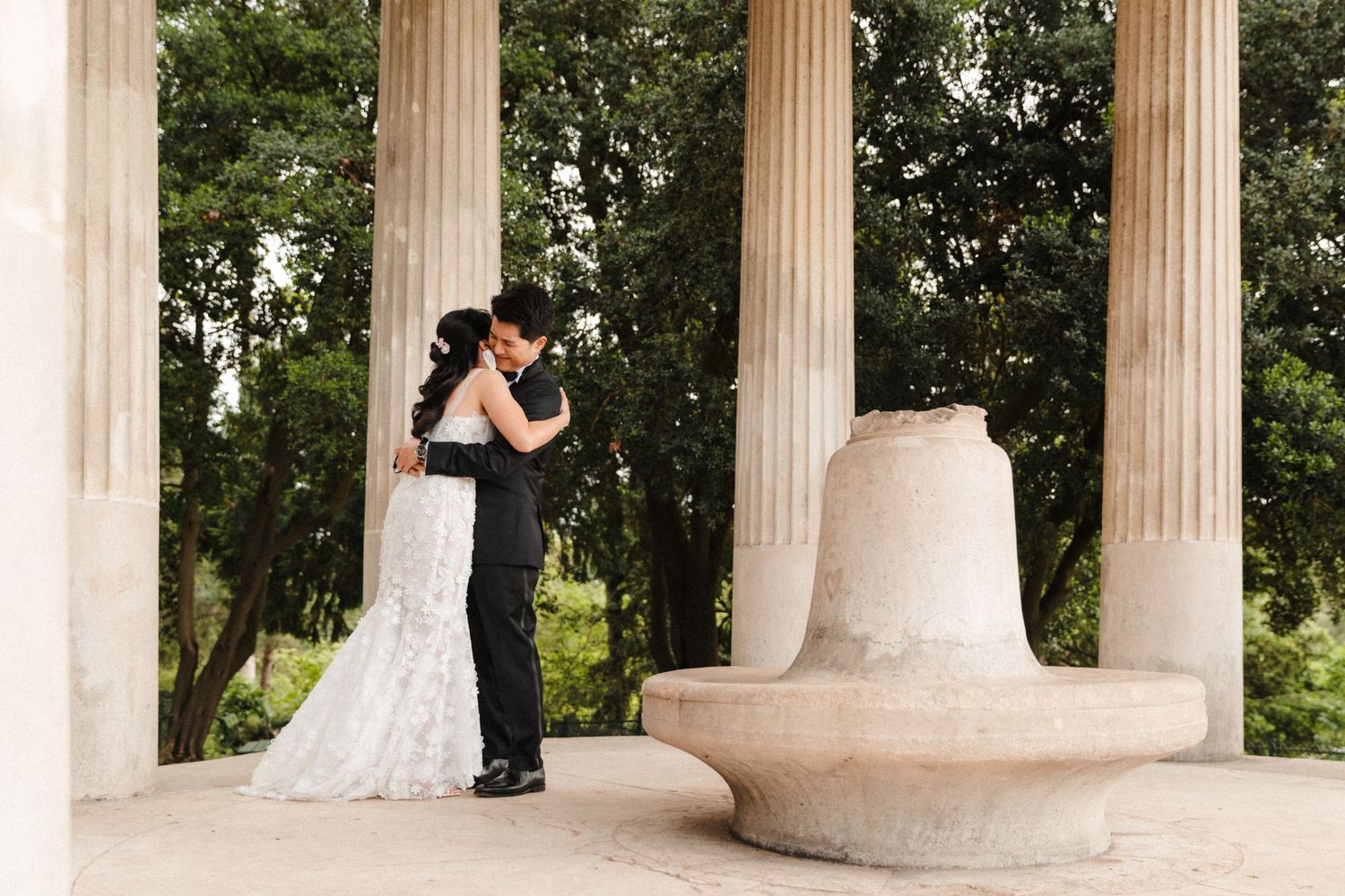 Bride and groom embrace among classical stone columns with lush green trees in background at elegant outdoor wedding venue.