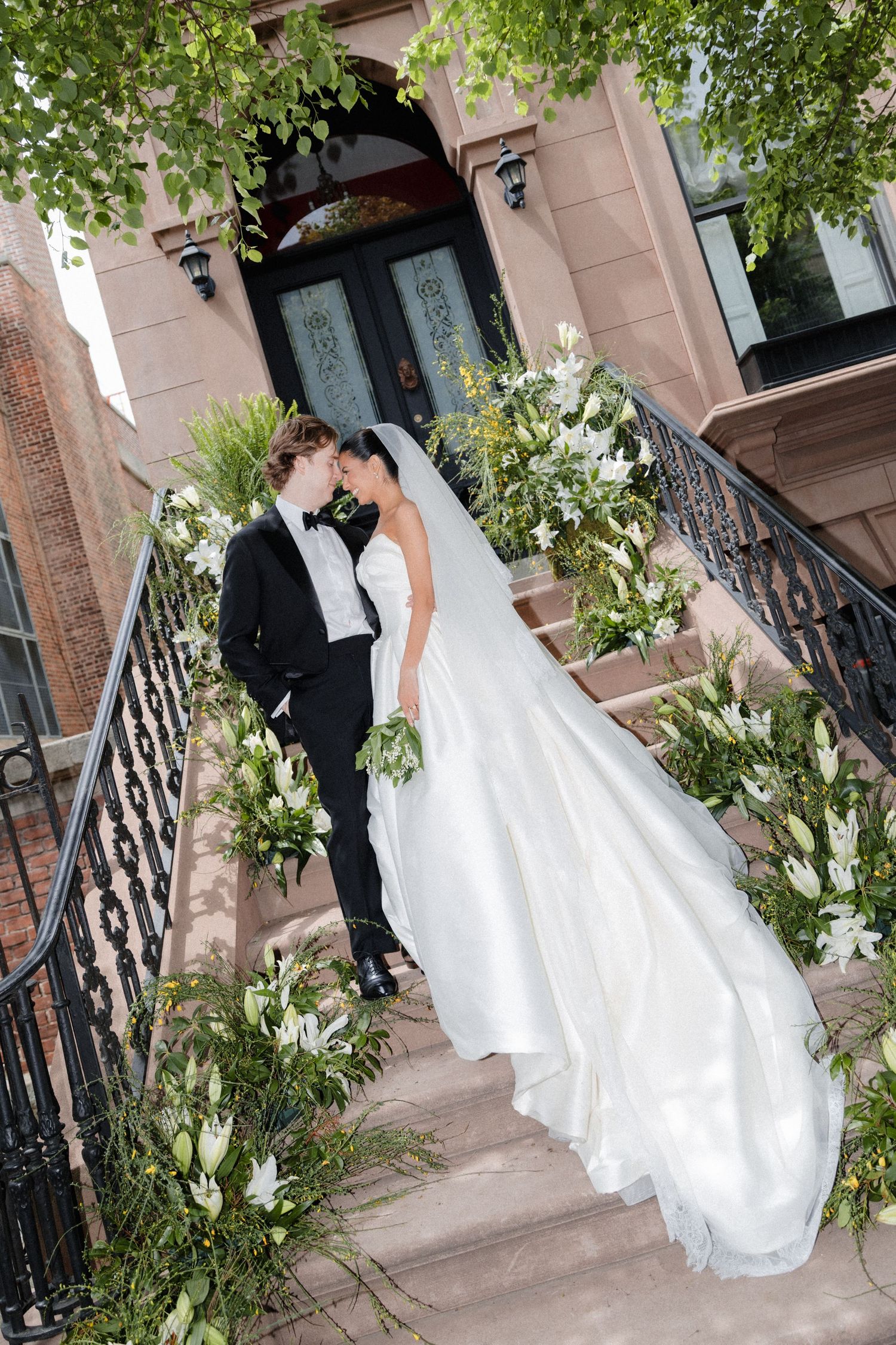 Bride and groom share a romantic moment on flower-adorned brownstone steps, surrounded by white lilies and lush greenery.