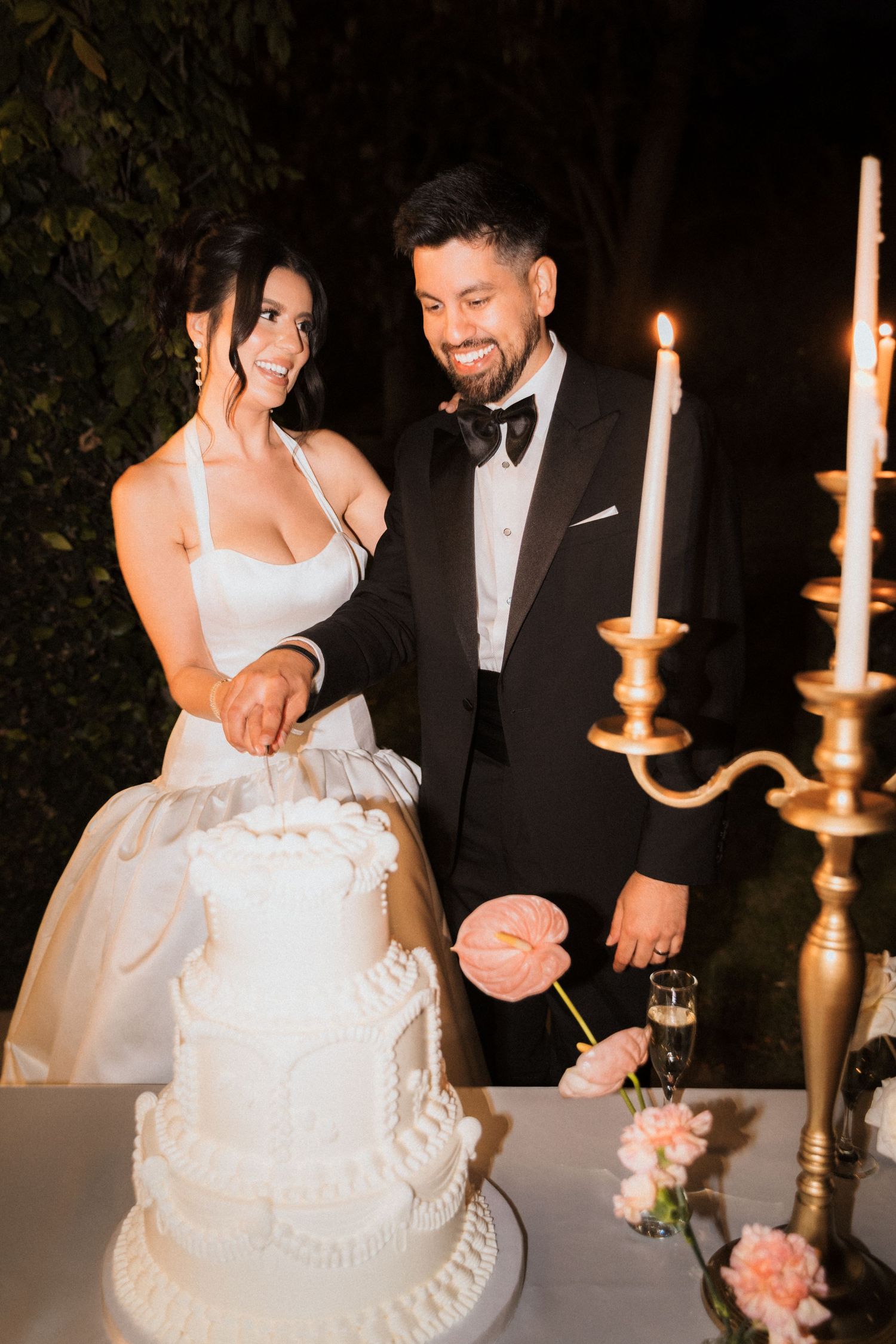A bride and groom cut into their elegant white wedding cake together while holding gold candlesticks in a romantic evening setting.