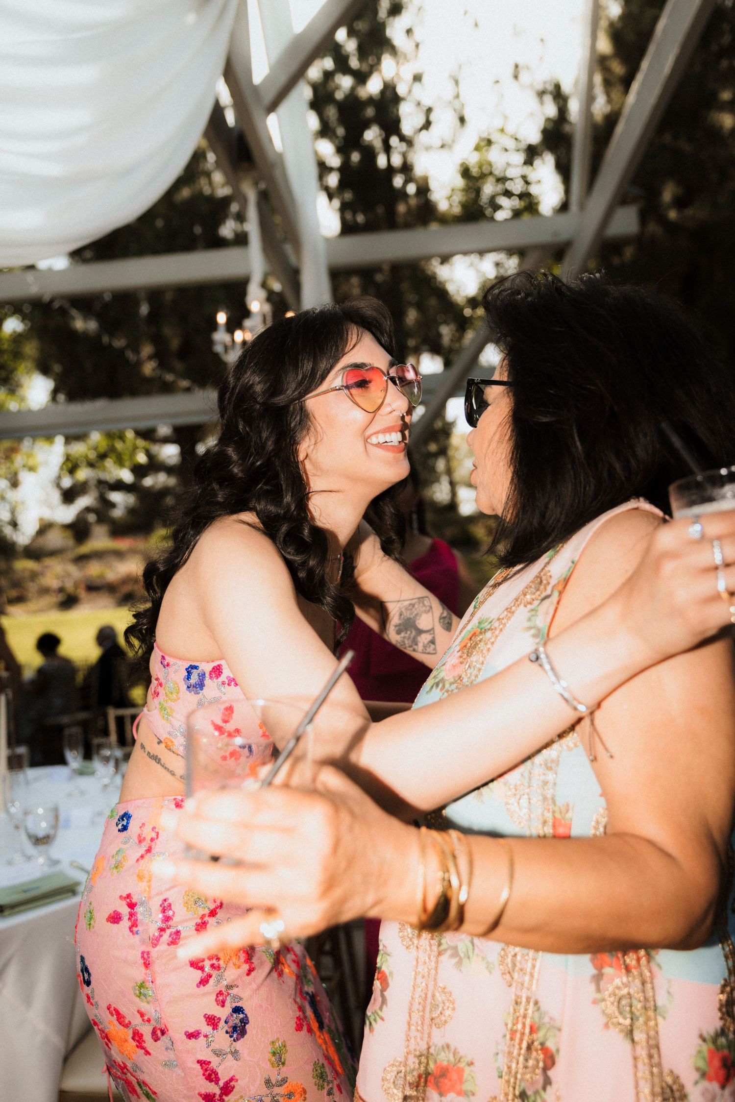 Two guests in floral dresses share a joyful moment dancing together under a white tent at an outdoor celebration.