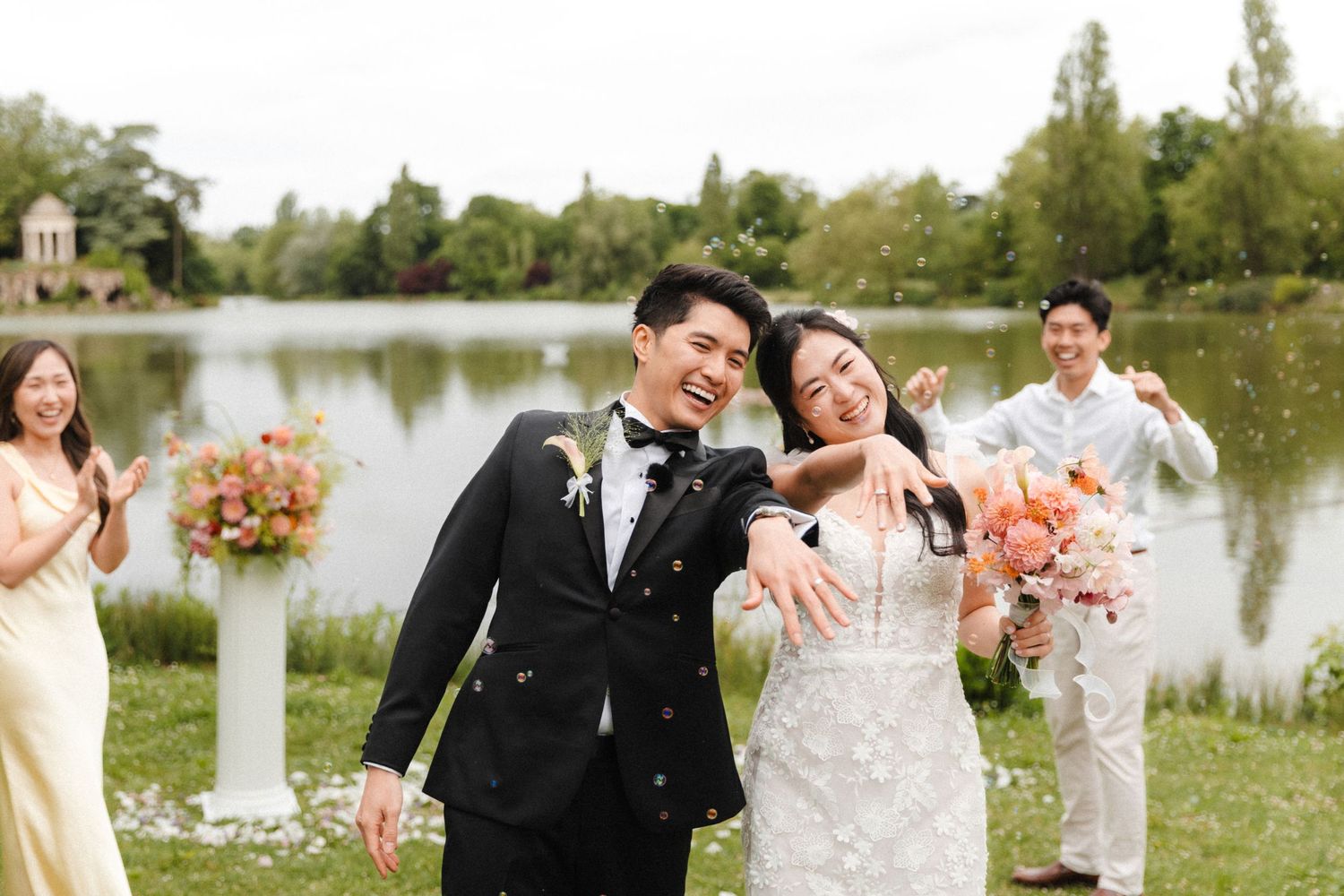 Happy Asian couple in wedding attire celebrating outdoors by a lake with guests throwing confetti.