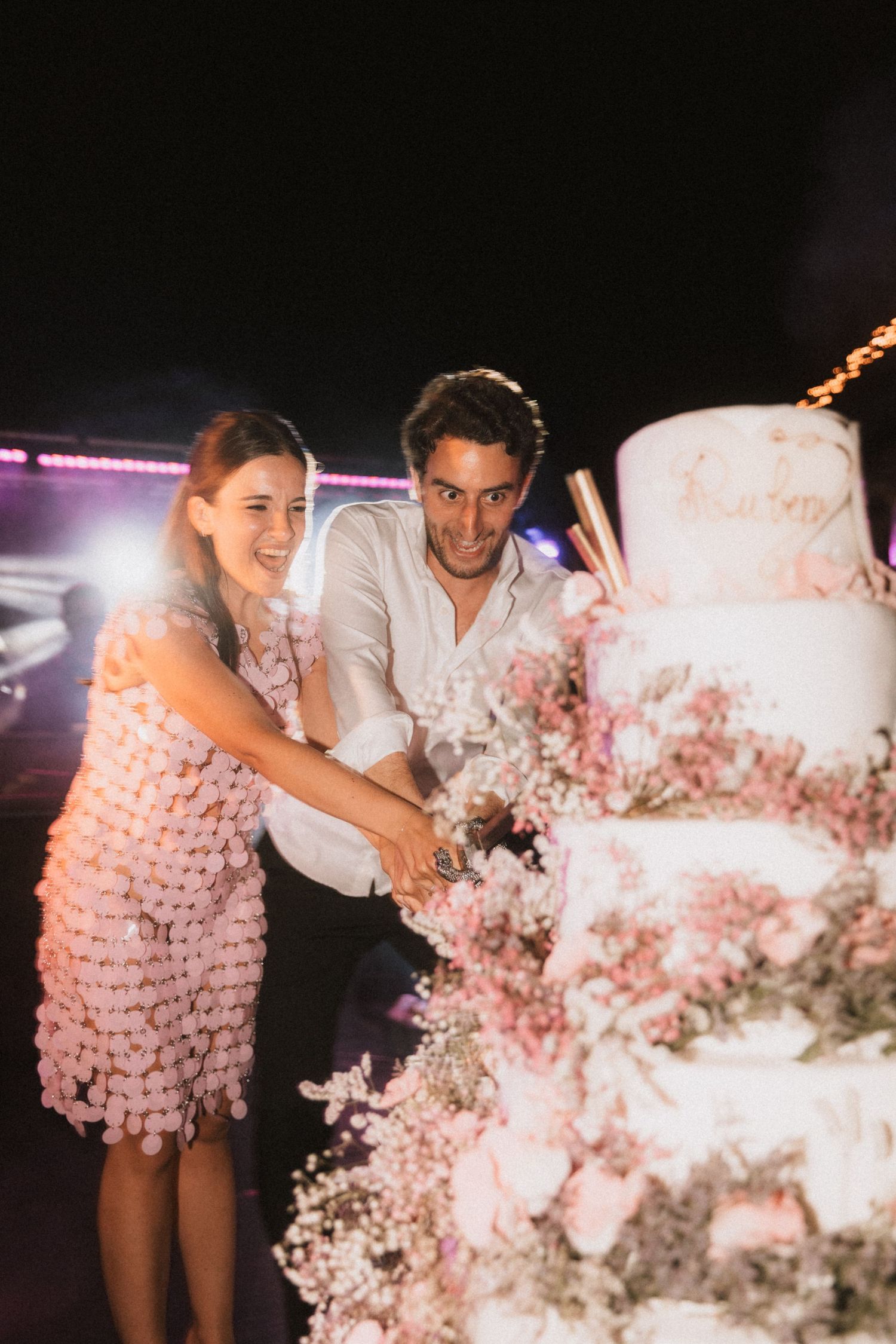 Wedding couple celebrates at reception near pink floral decorations and twinkling lights.