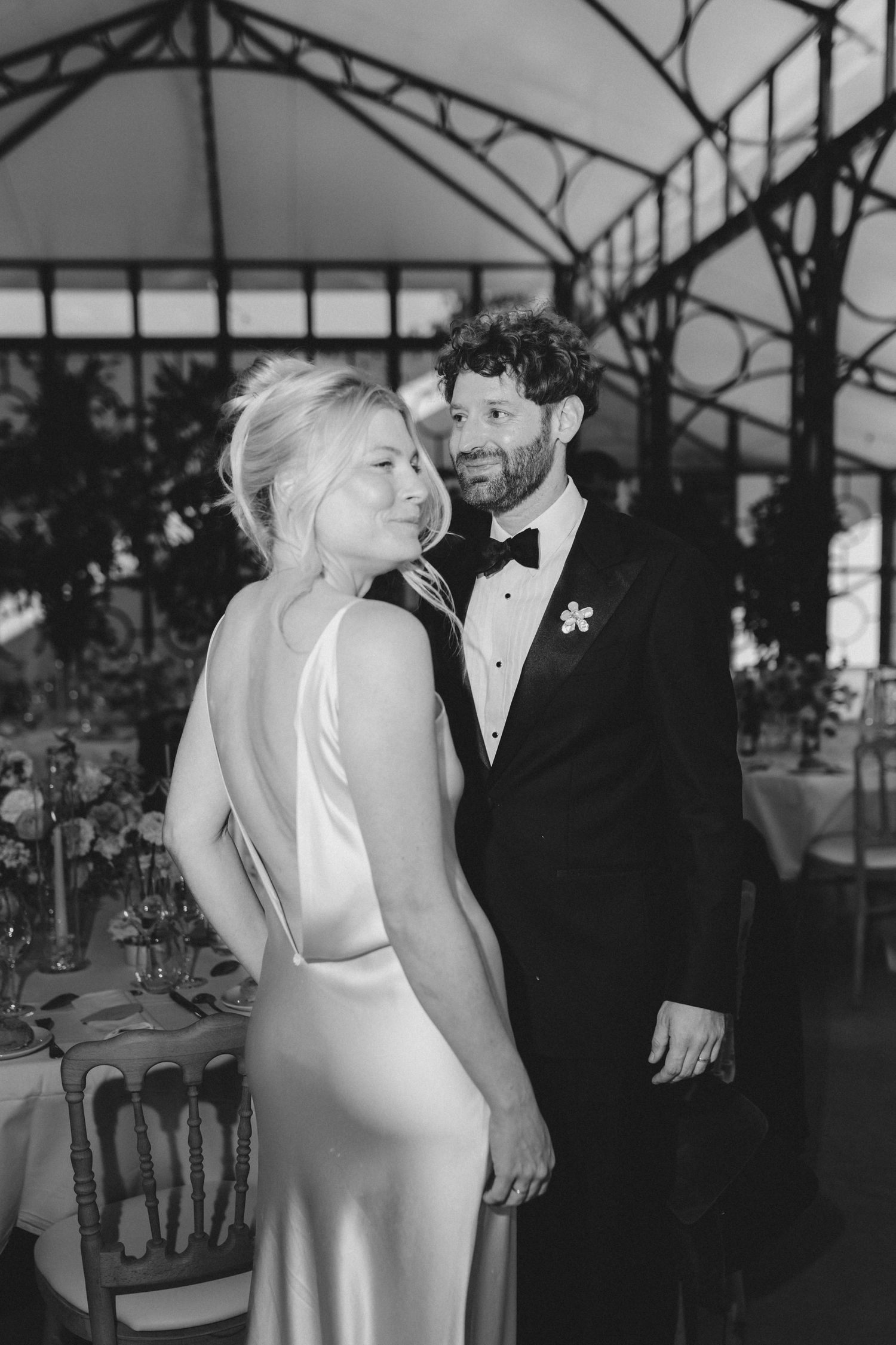 A romantic black and white wedding photo shows a couple embracing under an ornate glass ceiling with elegant decor.