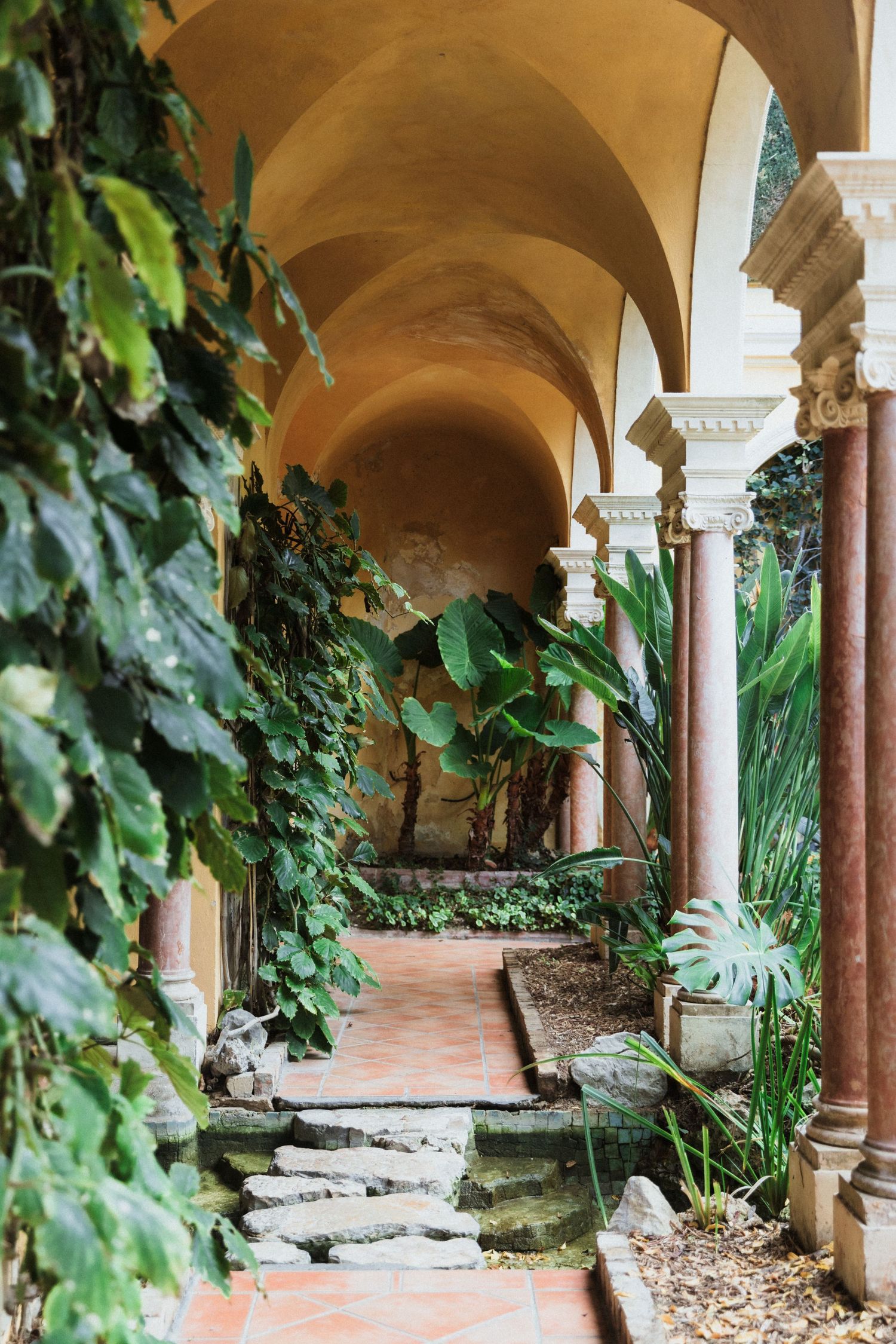 Villa Ephrussi de Rothschild - Lush tropical plants line an arched stone corridor  in a Mediterranean courtyard.