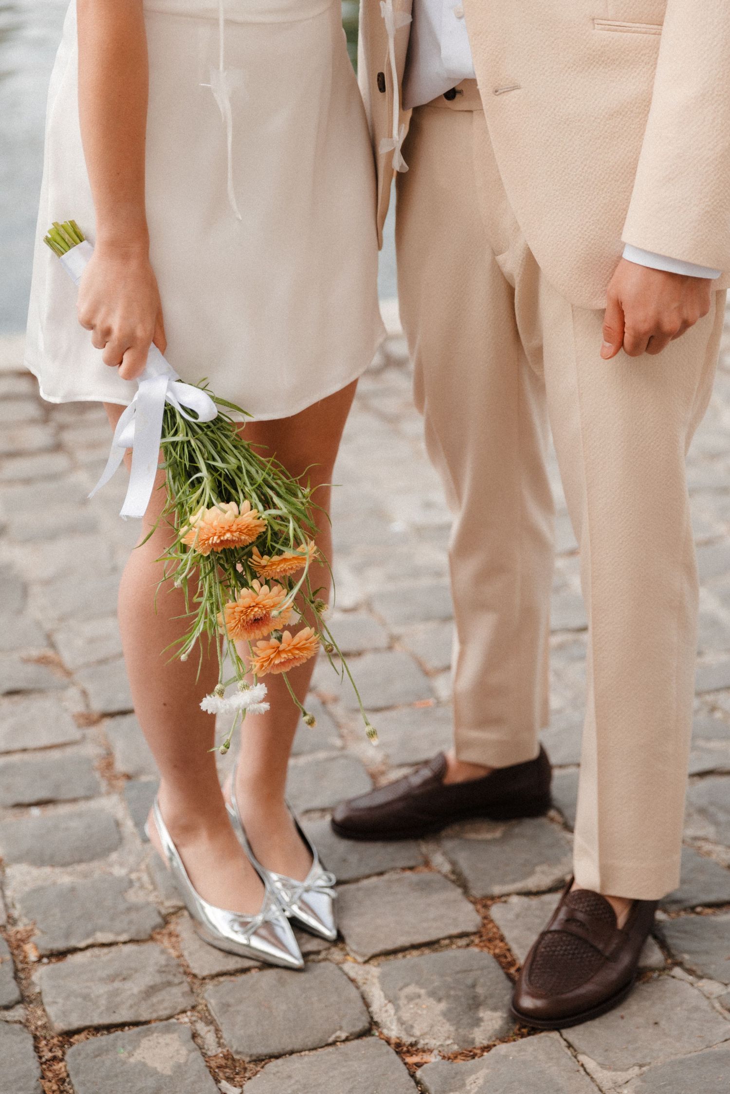 Bride in white dress with orange bouquet and silver shoes stands beside groom in beige suit on cobblestone.