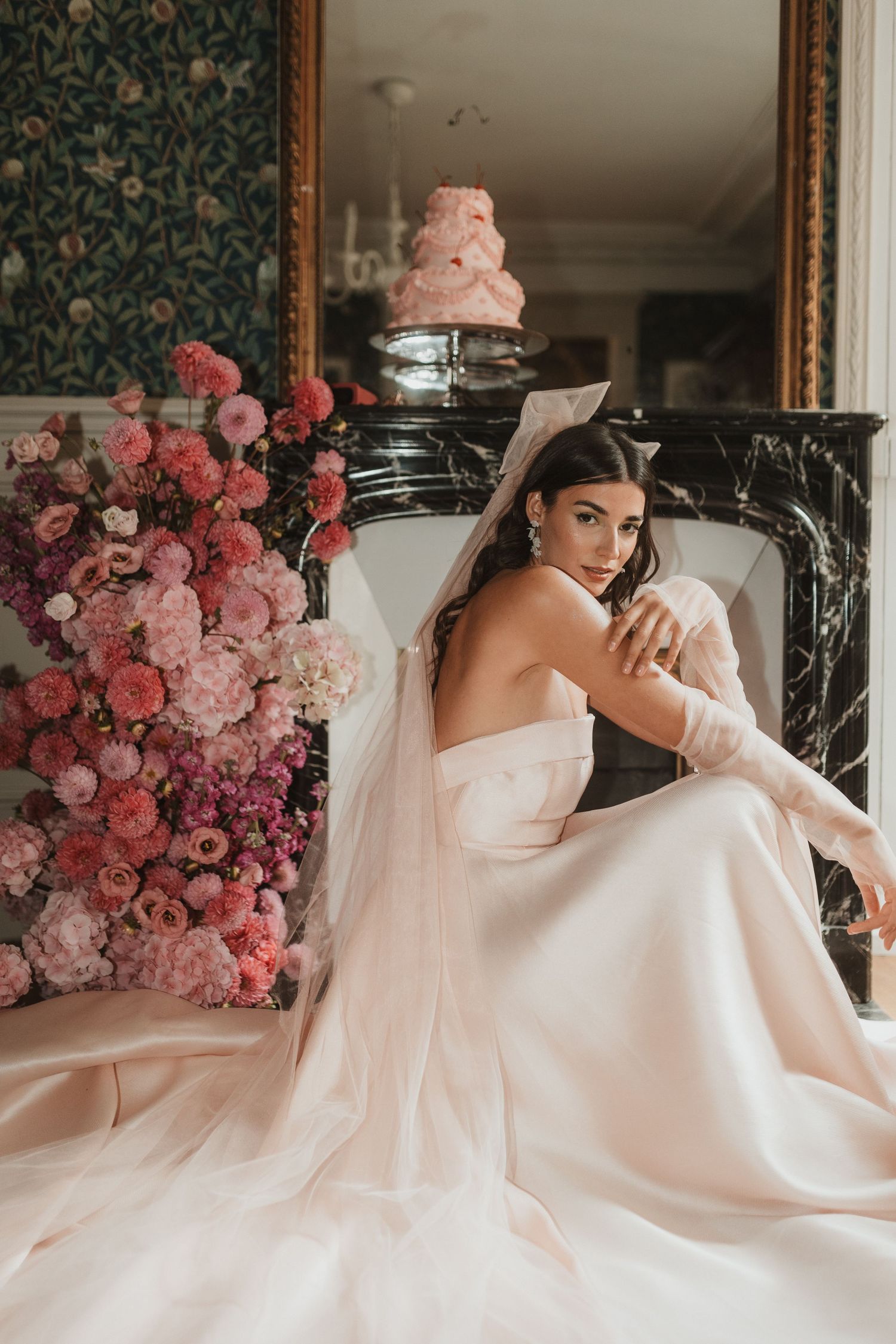 Bride in strapless blush gown with veil sits by marble fireplace, surrounded by lush pink floral arrangements and a tiered cake.
