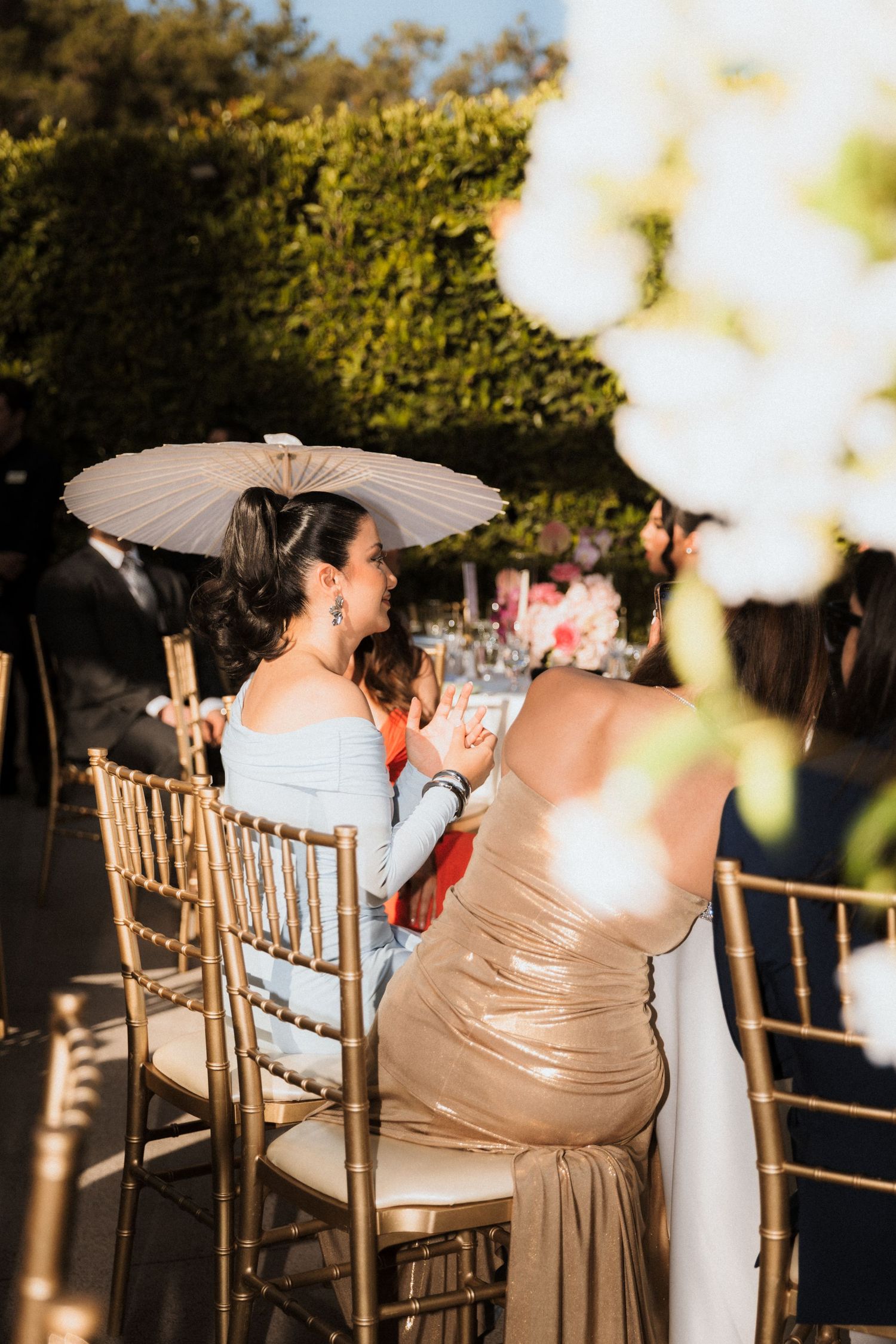 Wedding guests sit in wooden chairs holding parasols during an outdoor ceremony at sunset.