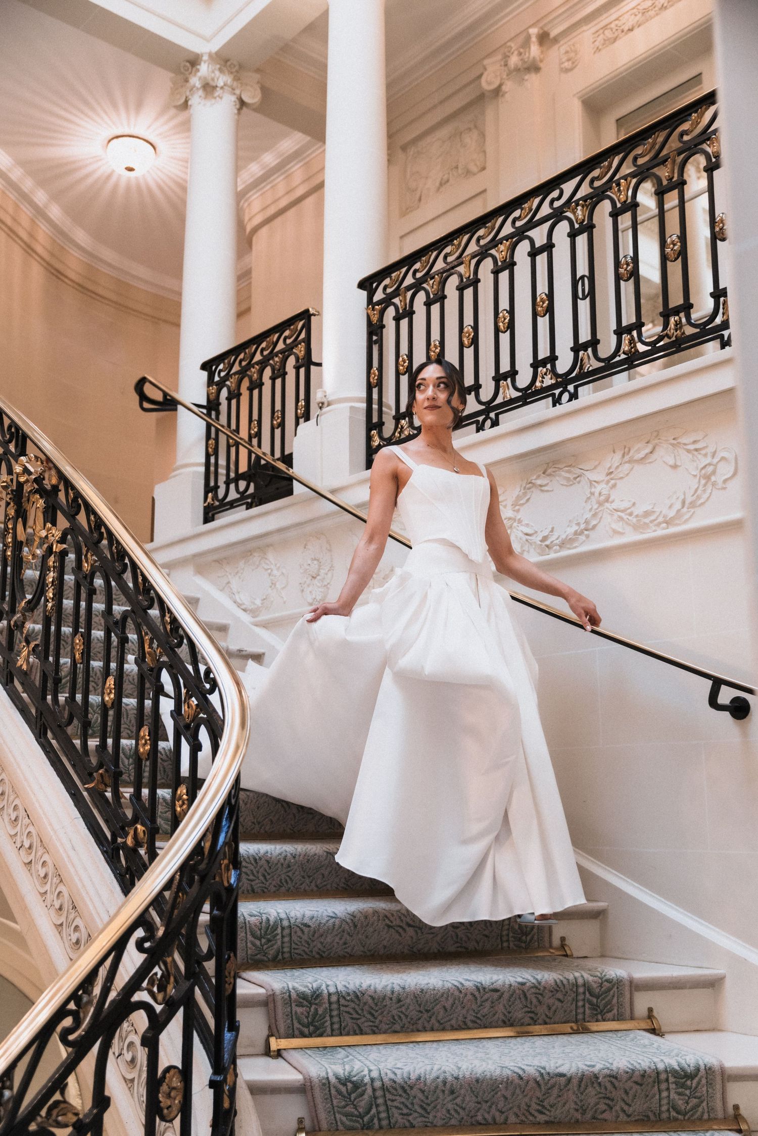 A bride in a white wedding gown poses elegantly on a grand staircase with ornate black railings and white columns.