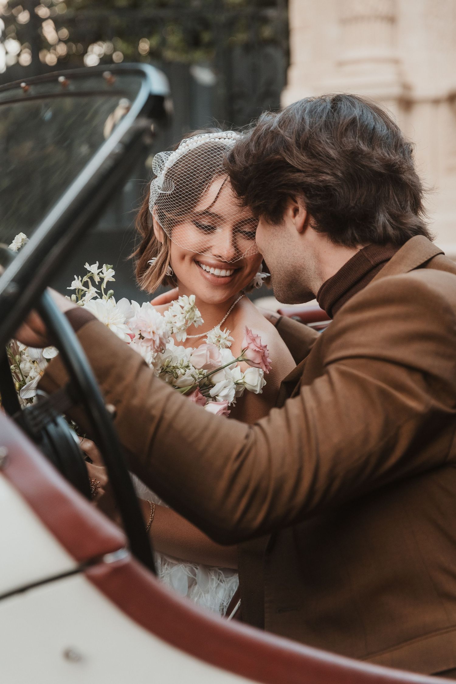 A smiling bride in a veil holds flowers as a groom in a brown suit leans in close beside a vintage convertible car.