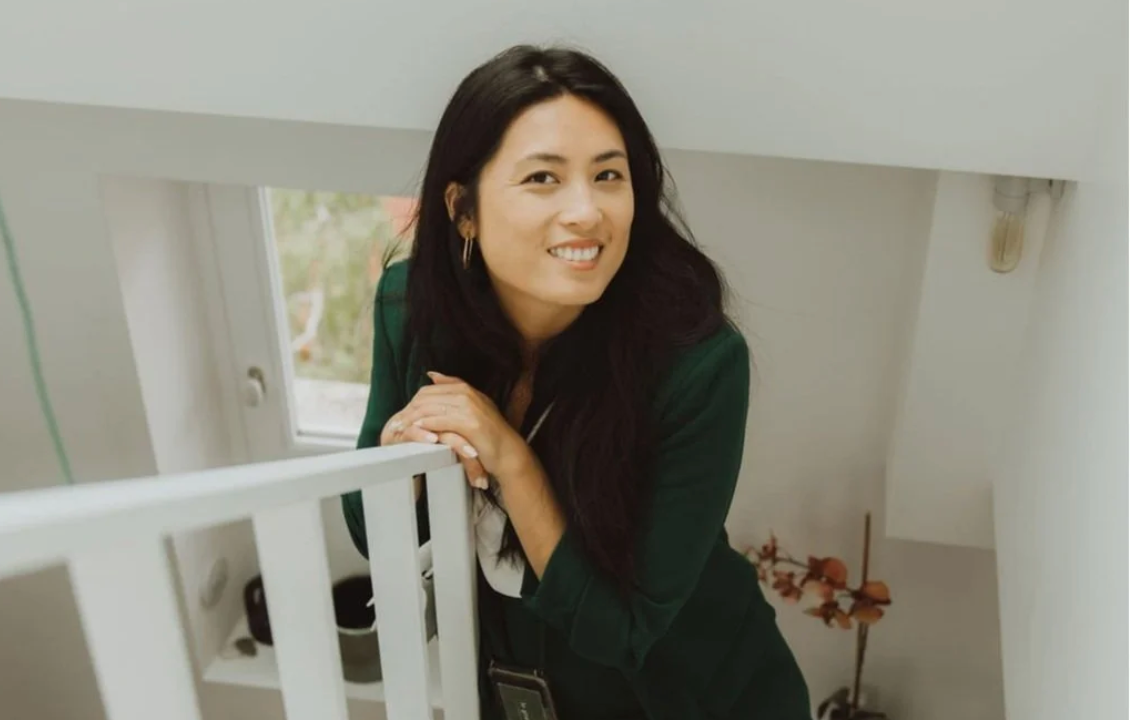 Smiling woman with long dark hair in a green blazer leans on a white stair railing in a bright, airy home with a window and flowers.