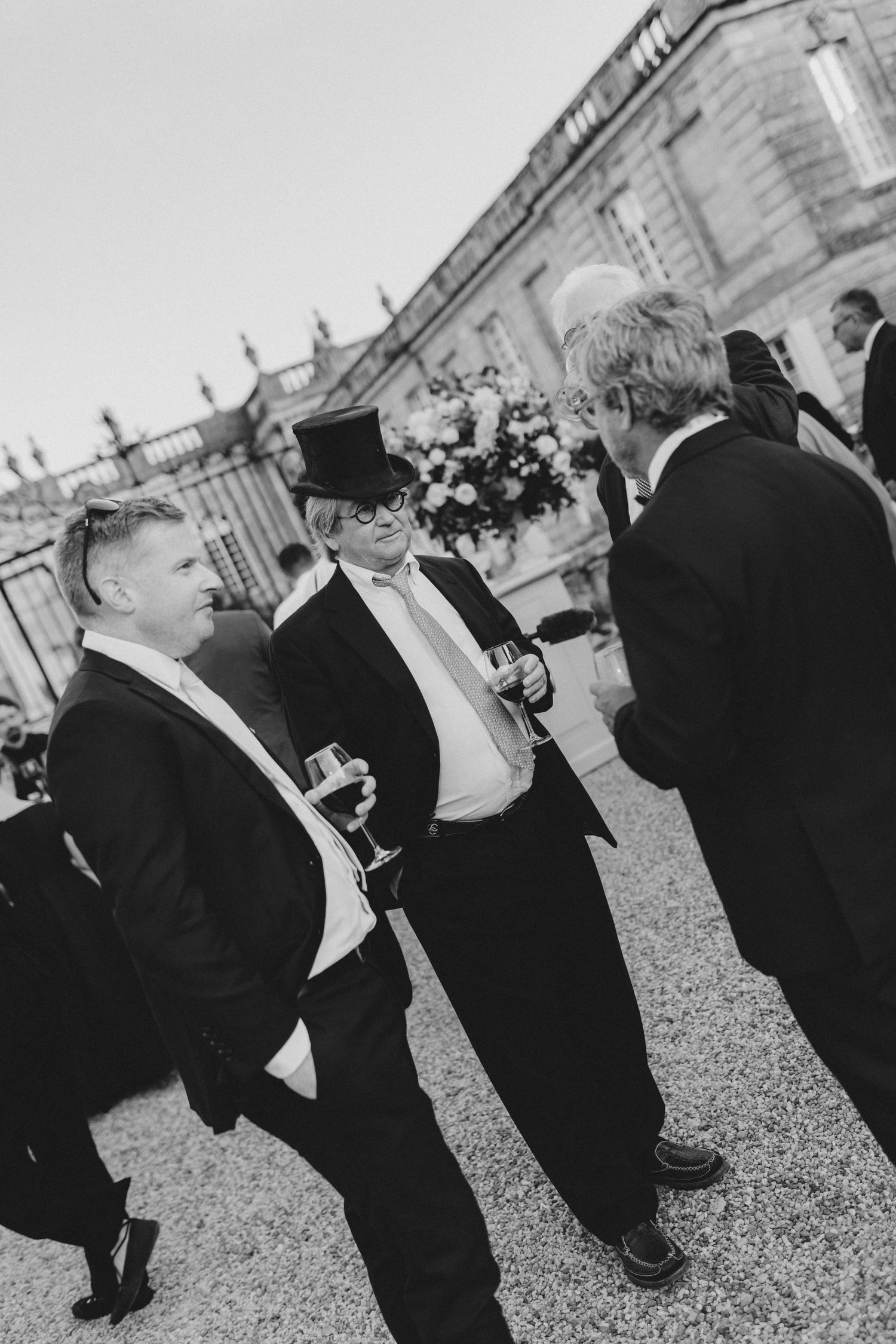 Three men in formal black tie attire and top hats chat during an elegant outdoor event at a grand venue.