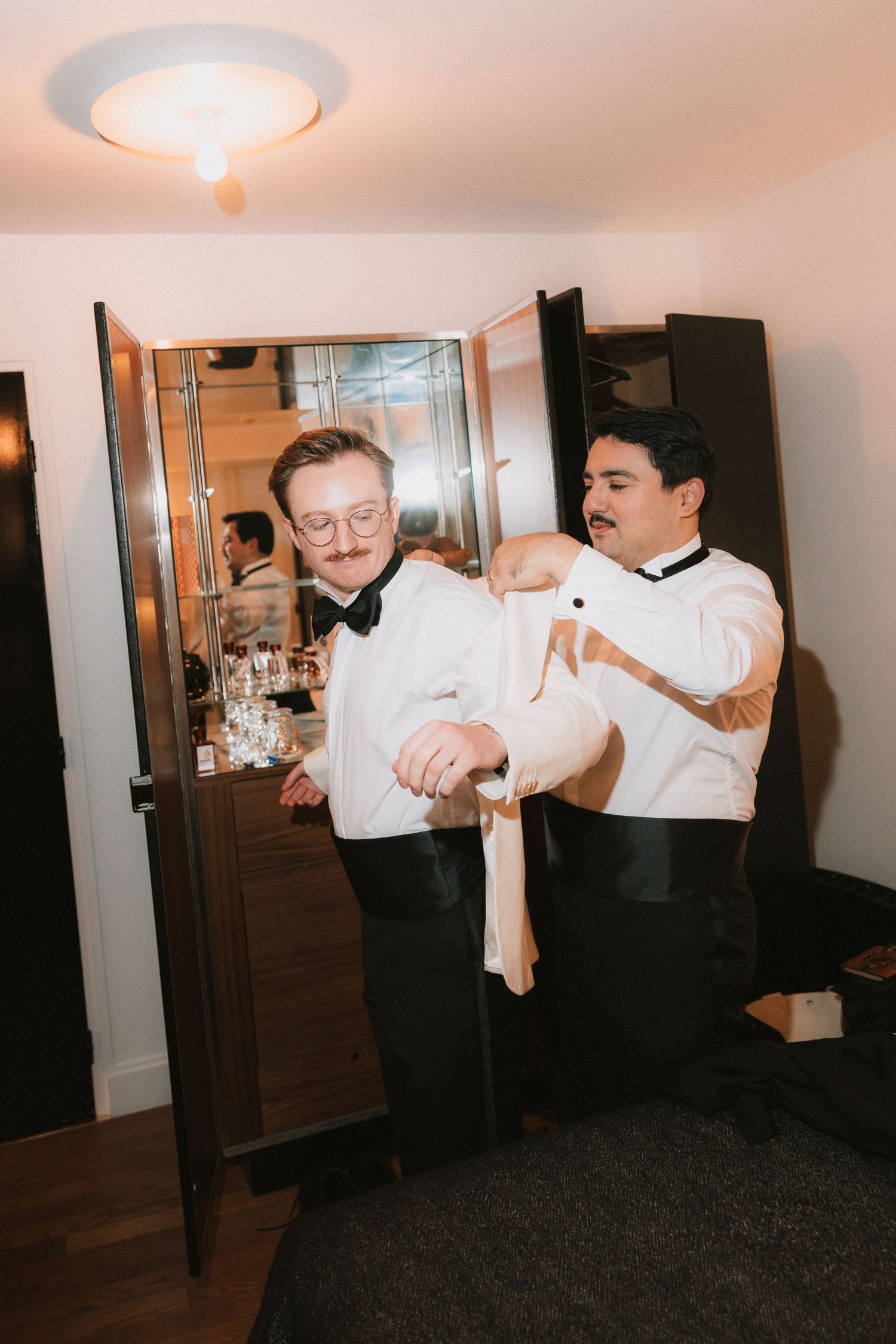 Two people in formal attire help each other get dressed, adjusting clothing in an indoor setting with a cabinet in background.