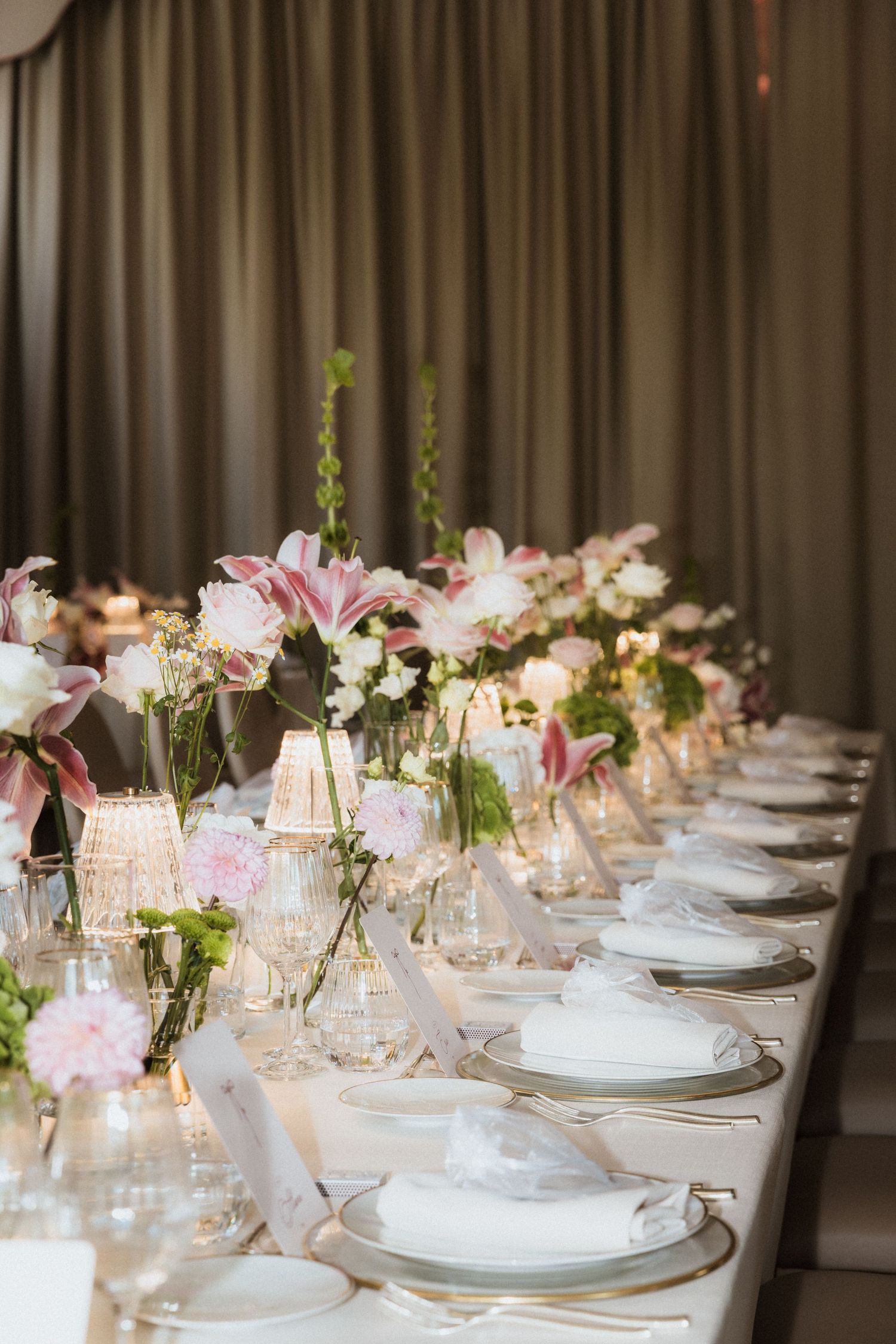 Elegant wedding reception table setting with pink lilies and white place settings in soft candlelight.