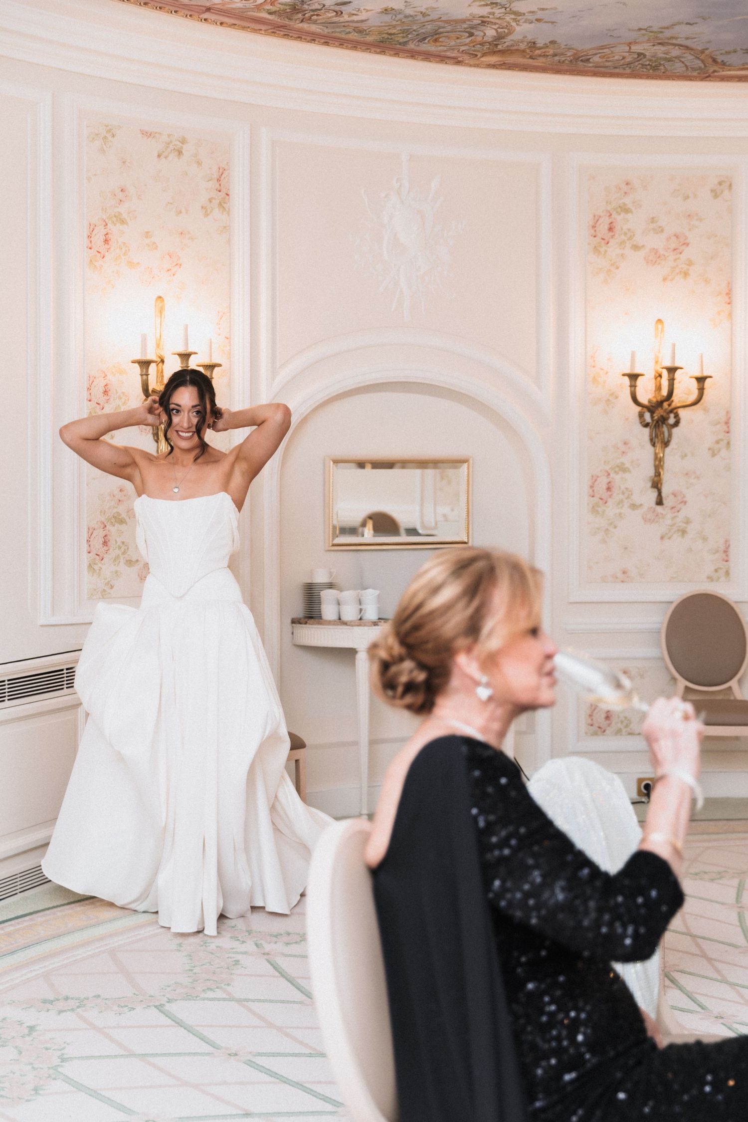 Two people in an elegant room with floral wallpaper, one wearing a white wedding dress and another in a black sequin gown.