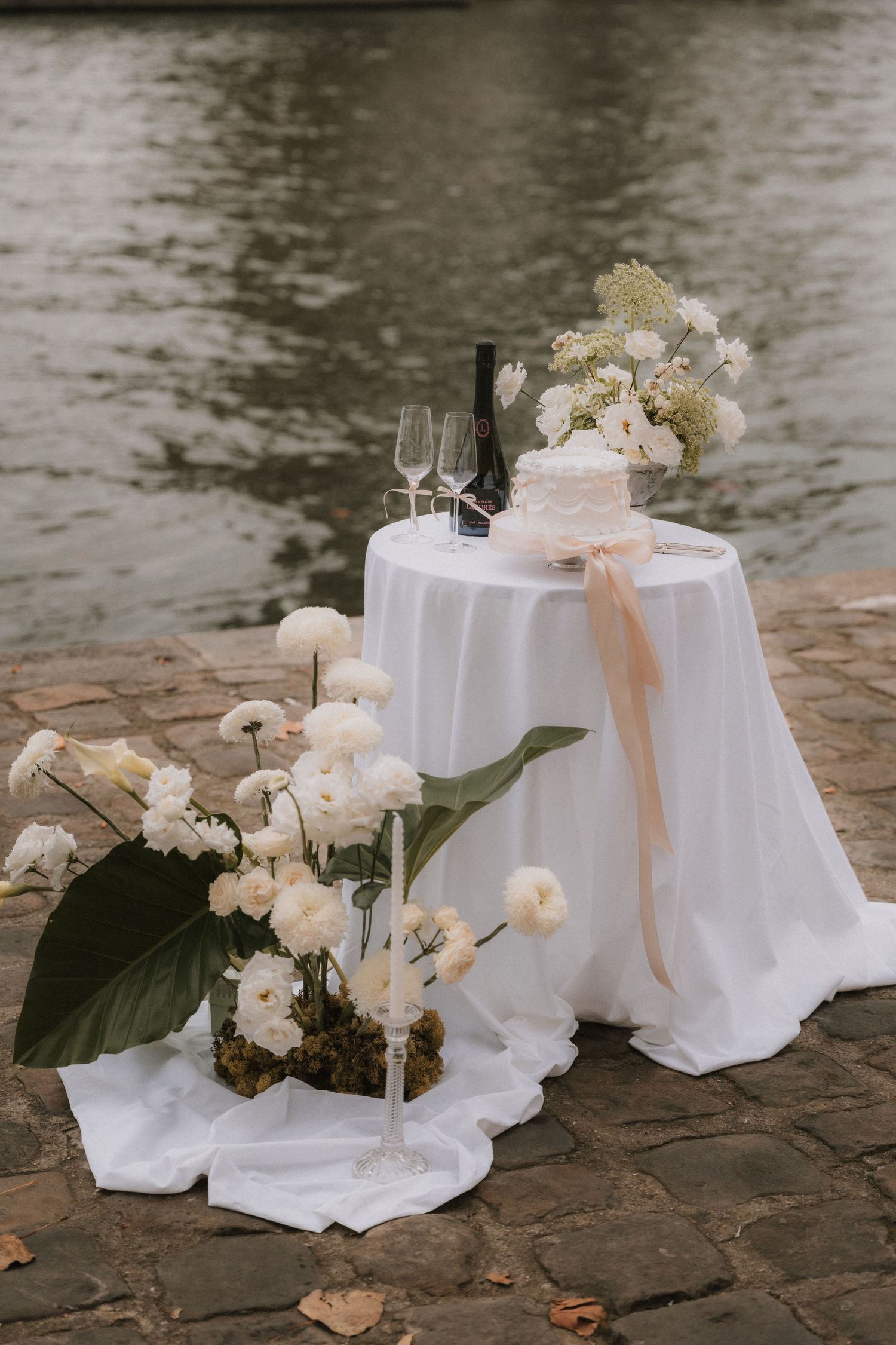 Elegant outdoor wedding setup with white cake, champagne flutes, and floral arrangements on a draped table by the water.