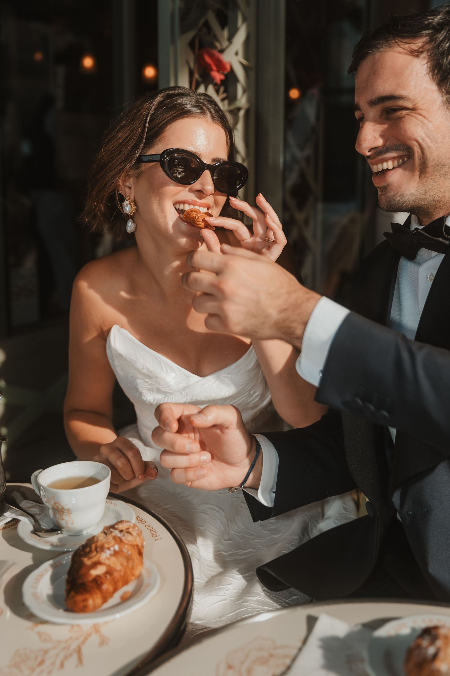 Bride and groom sharing a croissant at a cafe table, laughing together in elegant wedding attire.