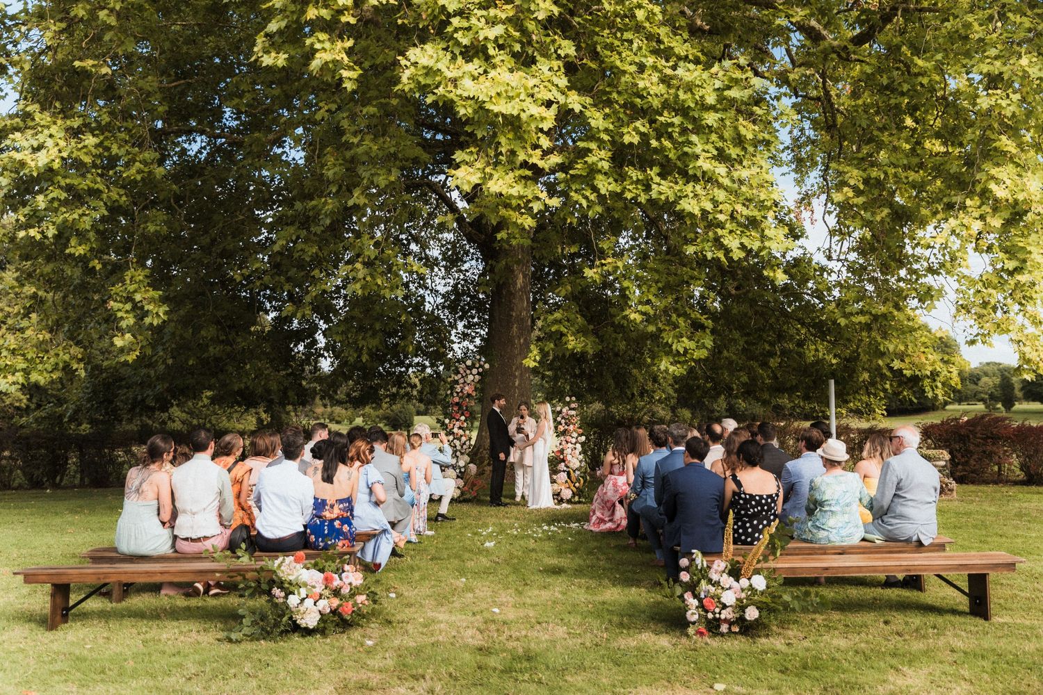 Outdoor wedding ceremony with guests seated on wooden benches under a large tree in a lush green garden.