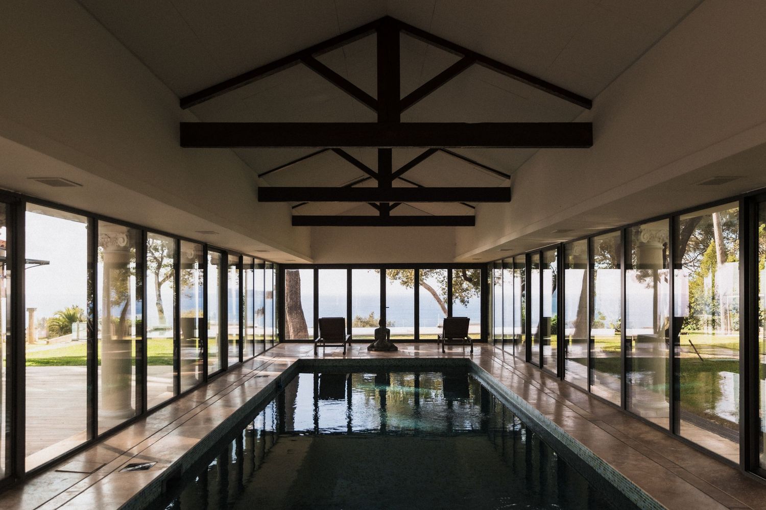 Villa Rocabella - Elegant indoor pool with vaulted ceiling, dark wood beams, and floor-to-ceiling windows overlooking a tropical waterfront.