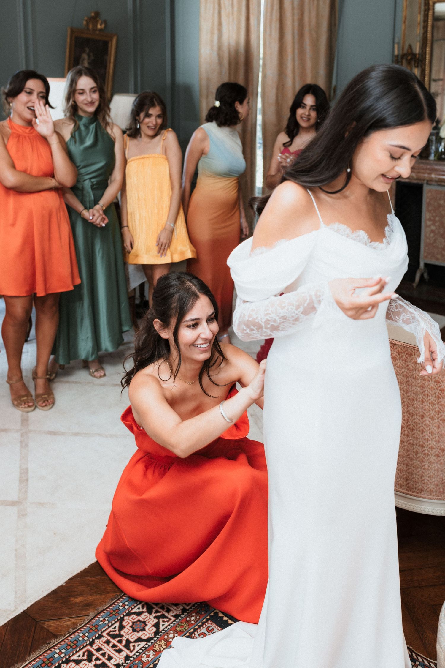 Bridesmaids in coral dresses help bride in white lace gown prepare for wedding ceremony.