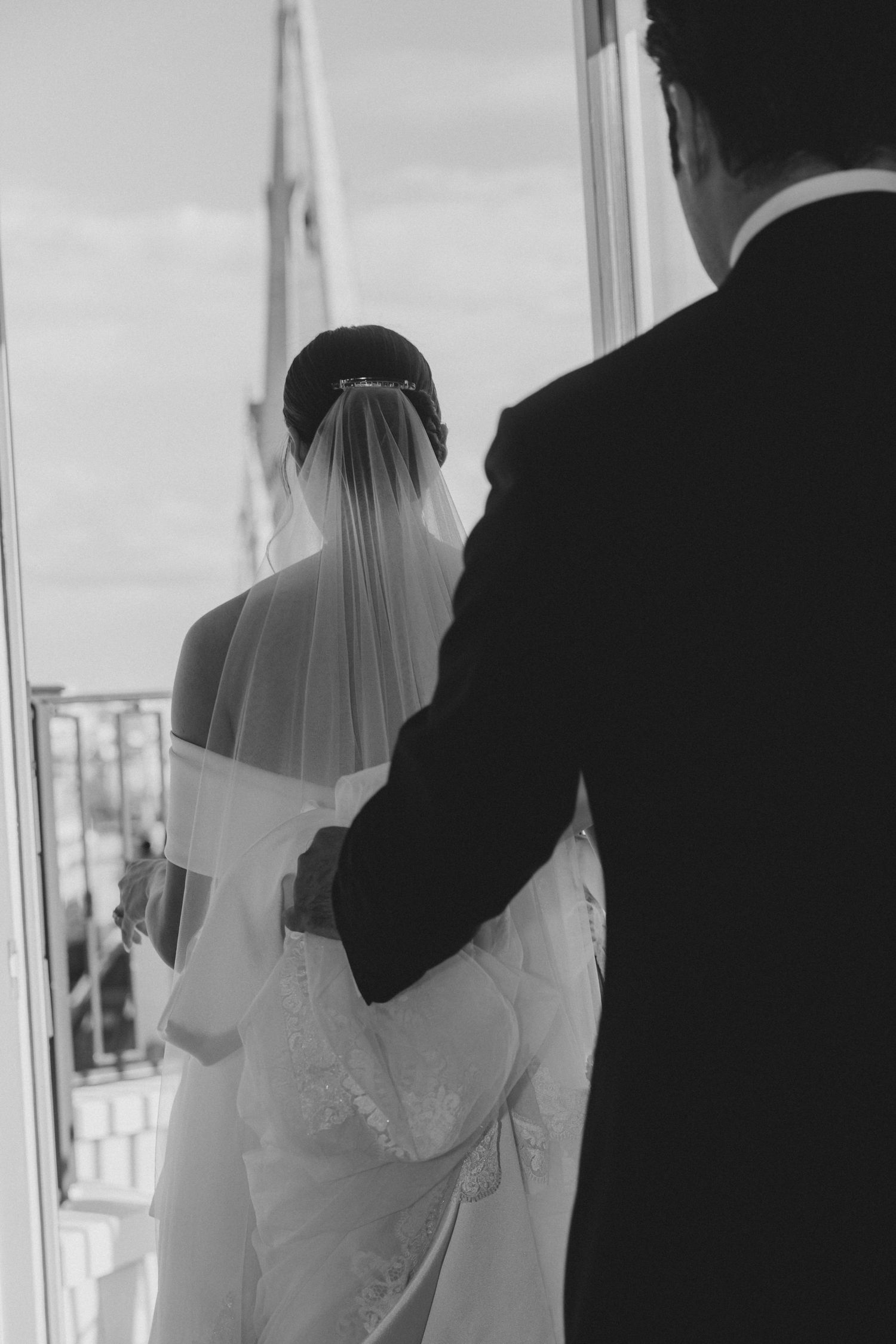 A bride and groom share an intimate moment by a window in black and white photography.