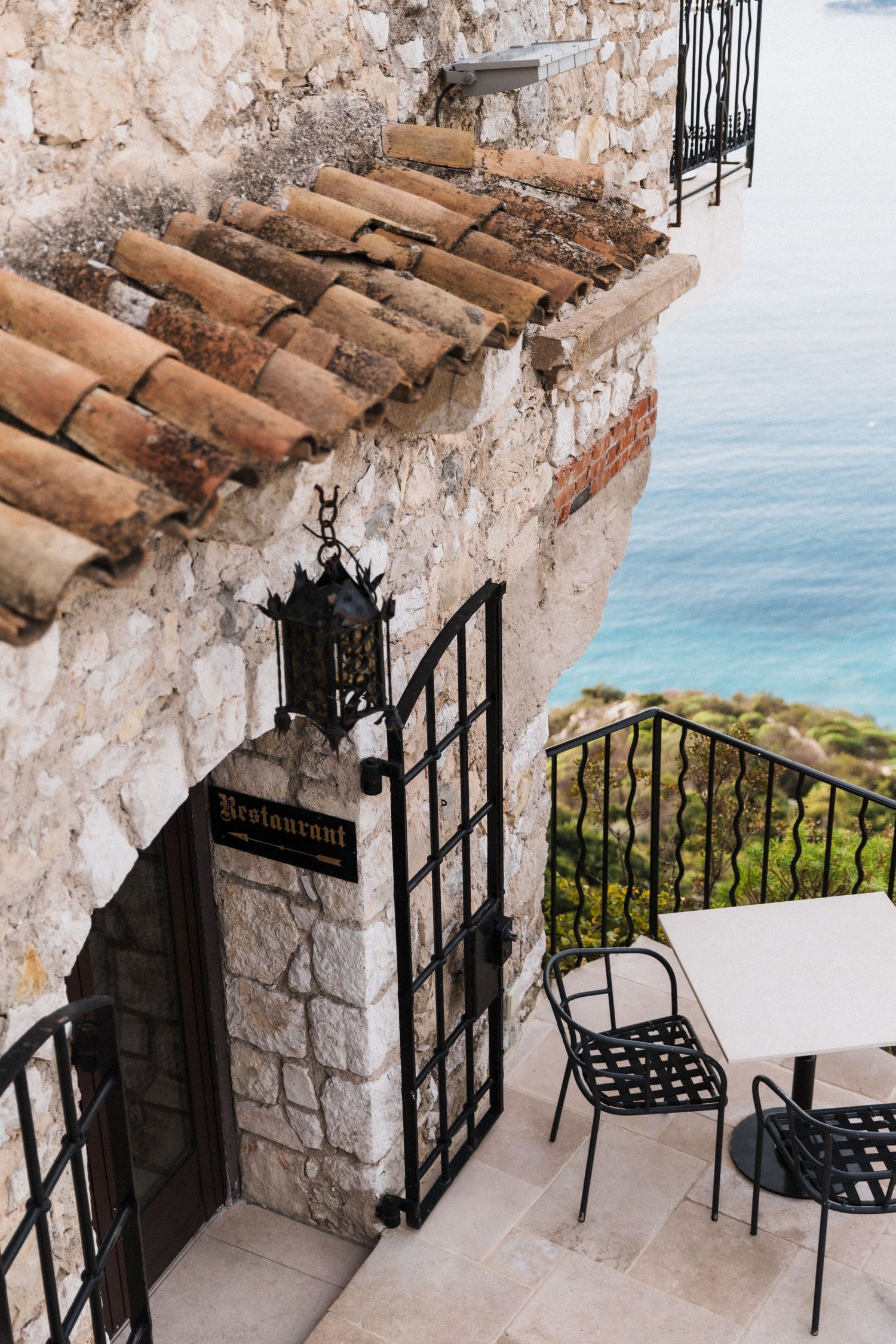 Château Eza - Stone restaurant entrance with wrought iron gate and terracotta roof tiles overlooking turquoise Mediterranean sea.