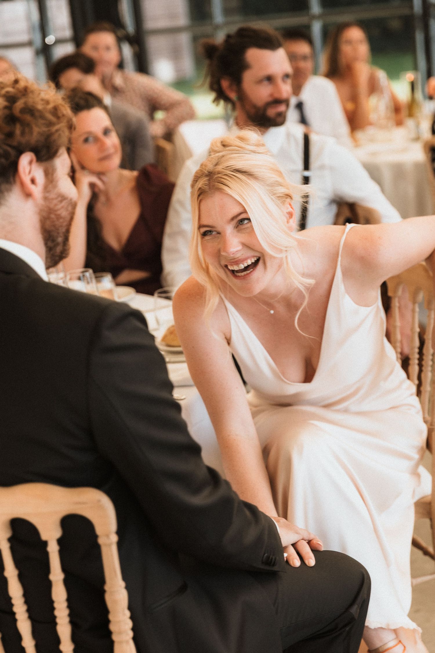 A laughing blonde bride in a white slip dress reaches out to touch a guest at an elegant wedding reception.