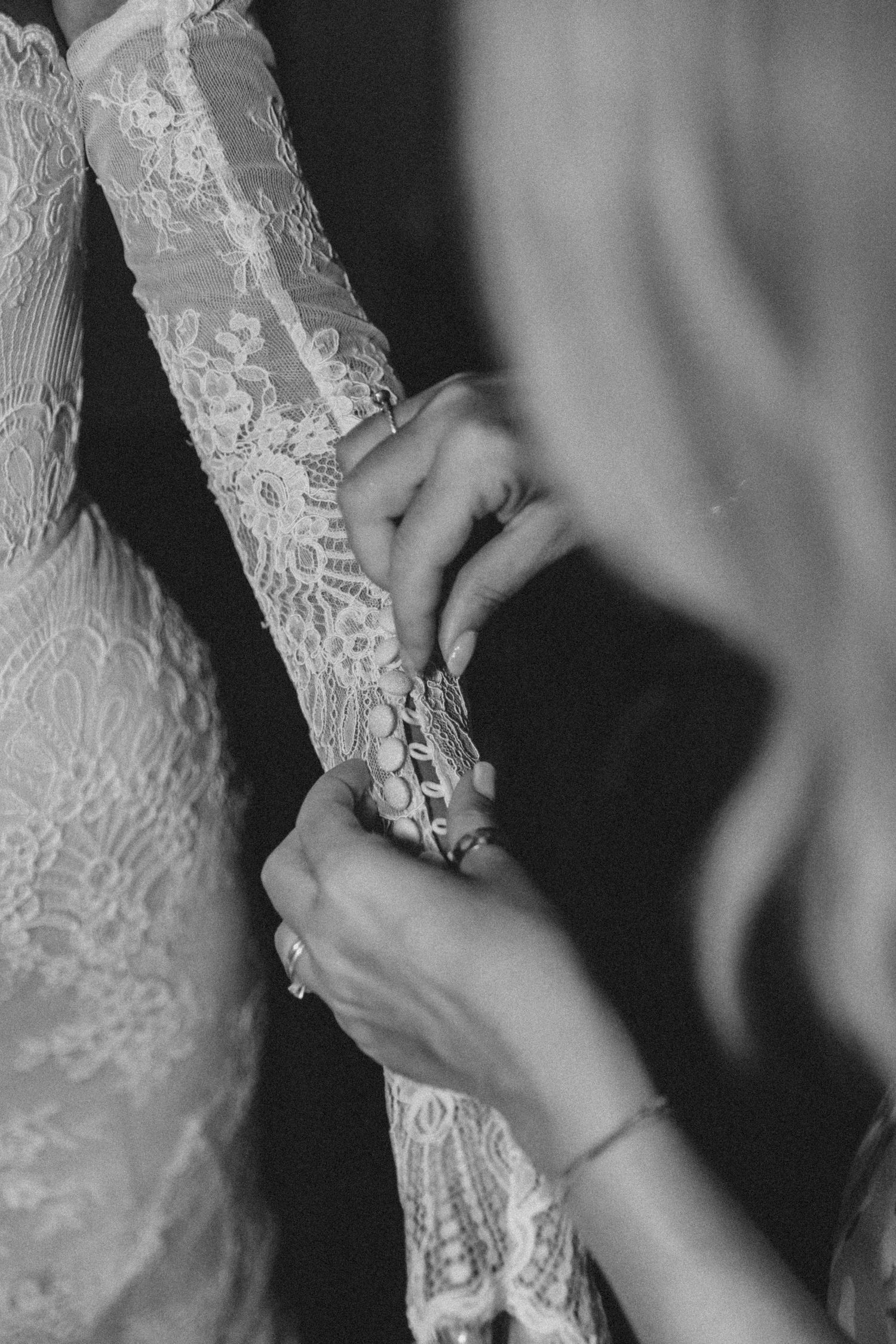 Close-up black and white image of hands buttoning up intricate lace details on a wedding dress.