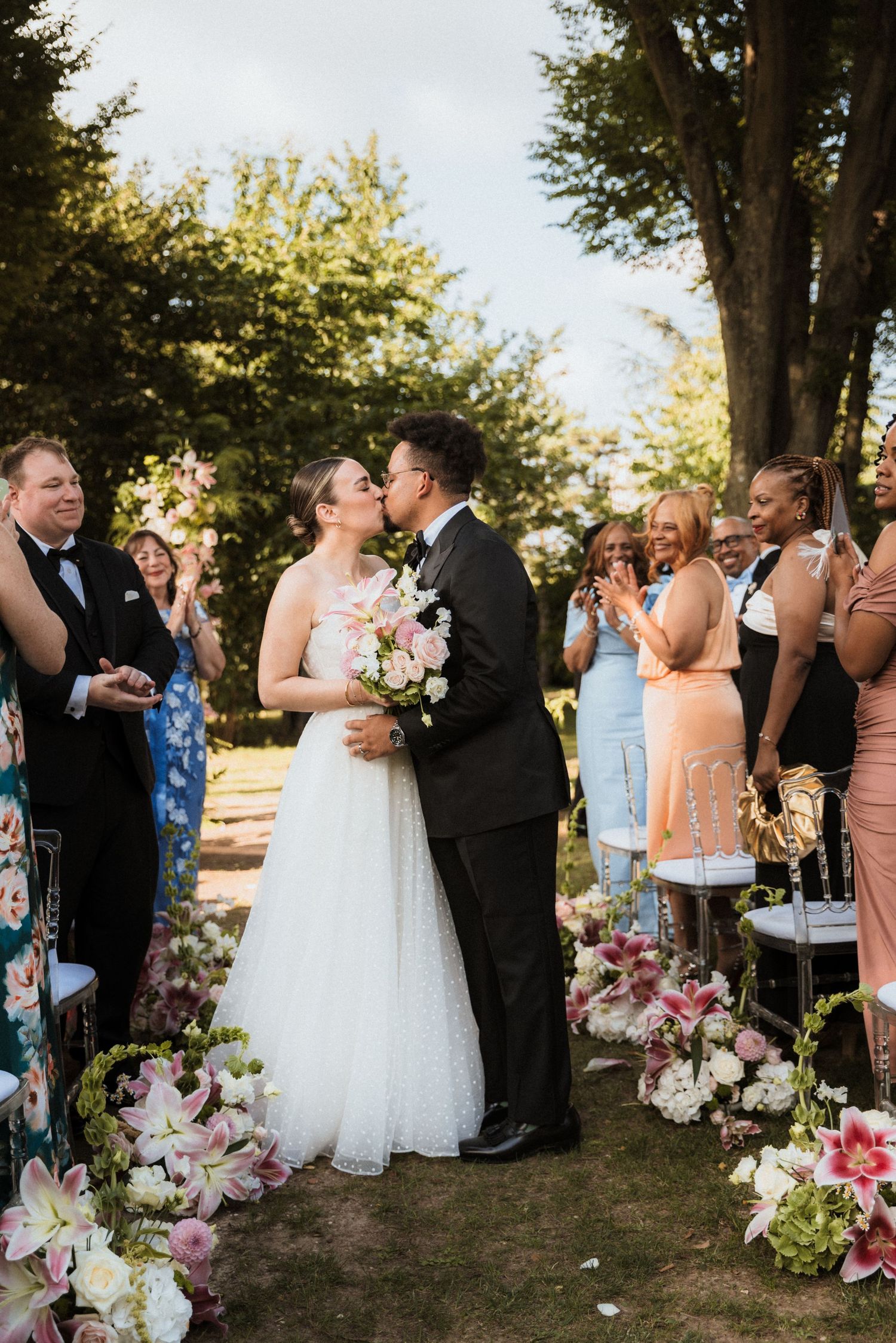 Wedding party and guests celebrating outdoors with floral arrangements and trees in background.