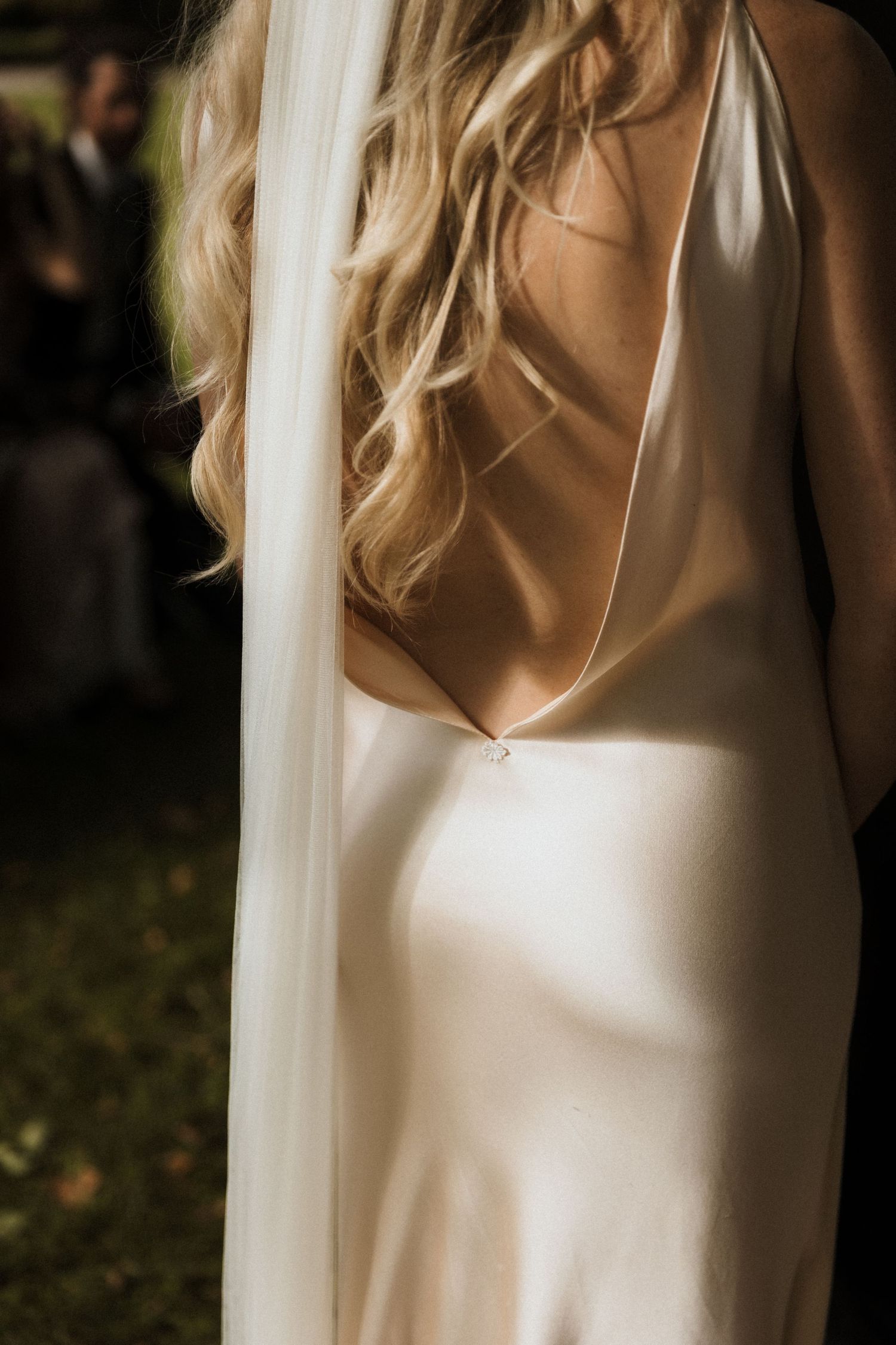 Bride in a backless ivory satin slip wedding gown with a flowing veil and wavy blonde hair.