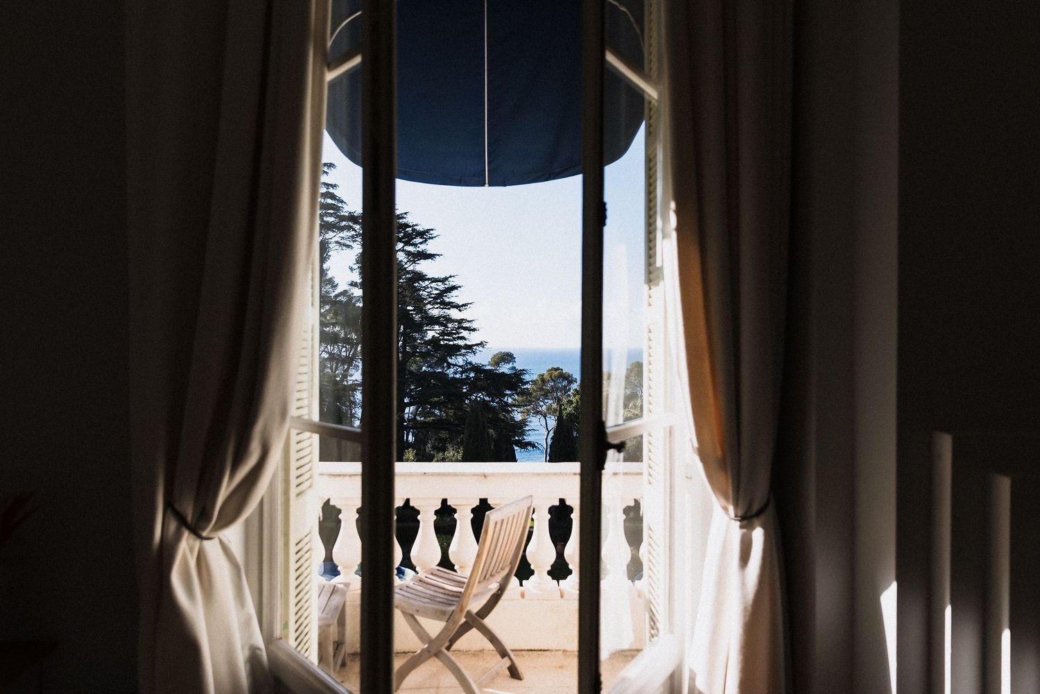 Villa Rocabella - Elegant French doors open to a sunlit balcony with a chair, ocean view, and lush trees framed by flowing curtains.