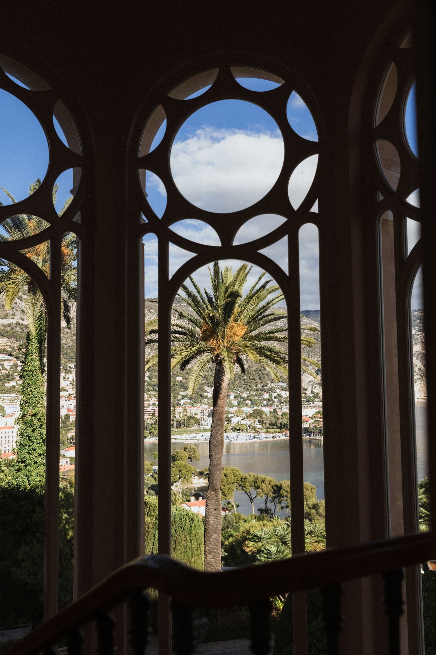 Villa Ephrussi de Rothschild - Ornate Gothic window frames a scenic Mediterranean coastal view with palm trees & blue water.