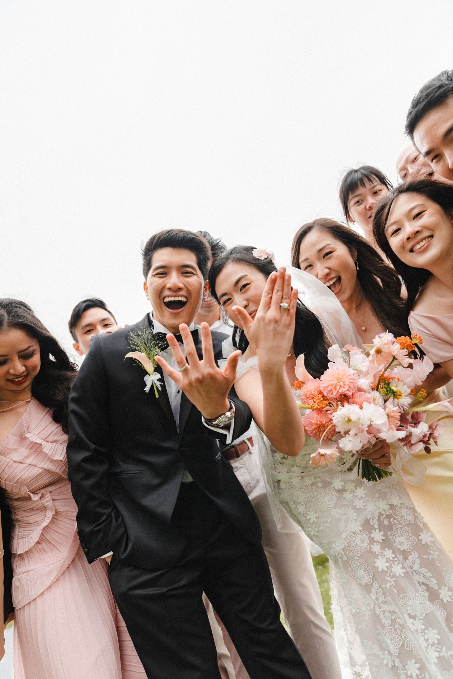 Joyful wedding party showing off rings, bride holding pink floral bouquet, groom in black suit, surrounded by smiling guests.